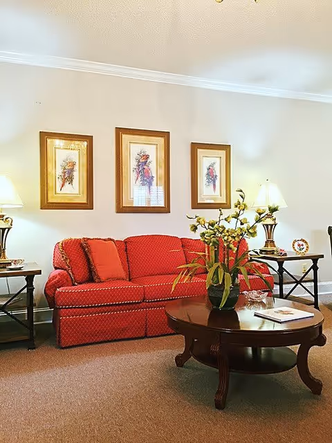 A cozy living room with a red patterned sofa adorned with matching cushions. Above the sofa, there are three framed floral artworks hanging on a light-colored wall. On either side of the sofa, there are dark wooden side tables with lamps and decorative items. In front of the sofa, a round wooden coffee table holds a vase with flowers and a book. The room has a warm and inviting atmosphere.