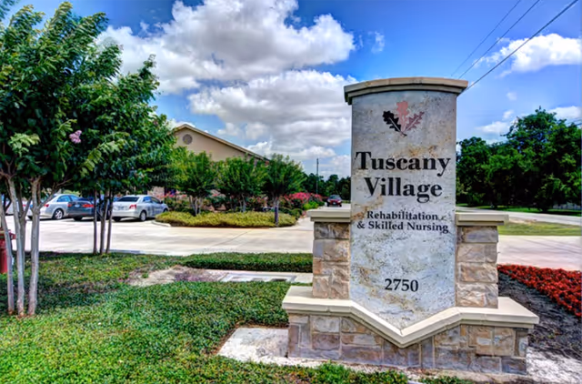 Stone sign for Tuscany Village Rehabilitation & Skilled Nursing with the address 2750, surrounded by green grass, trees, and a parking lot with cars under a partly cloudy blue sky.
