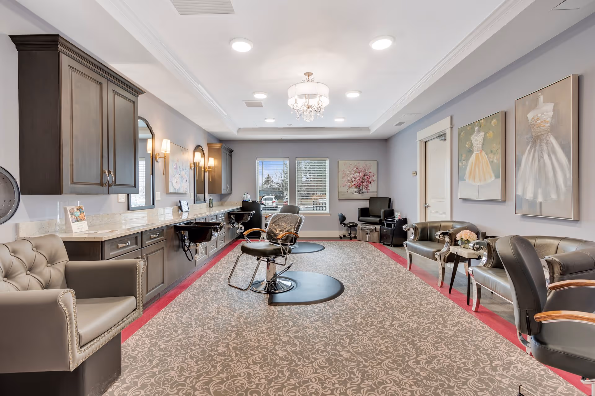 A spacious and well-lit salon area in a senior living facility with multiple styling chairs, mirrors, and sinks along one wall. The room features elegant decor including framed artwork of dresses on the walls, comfortable seating with armchairs, a patterned carpet, and a chandelier hanging from the ceiling. Large windows at the far end allow natural light to enter.