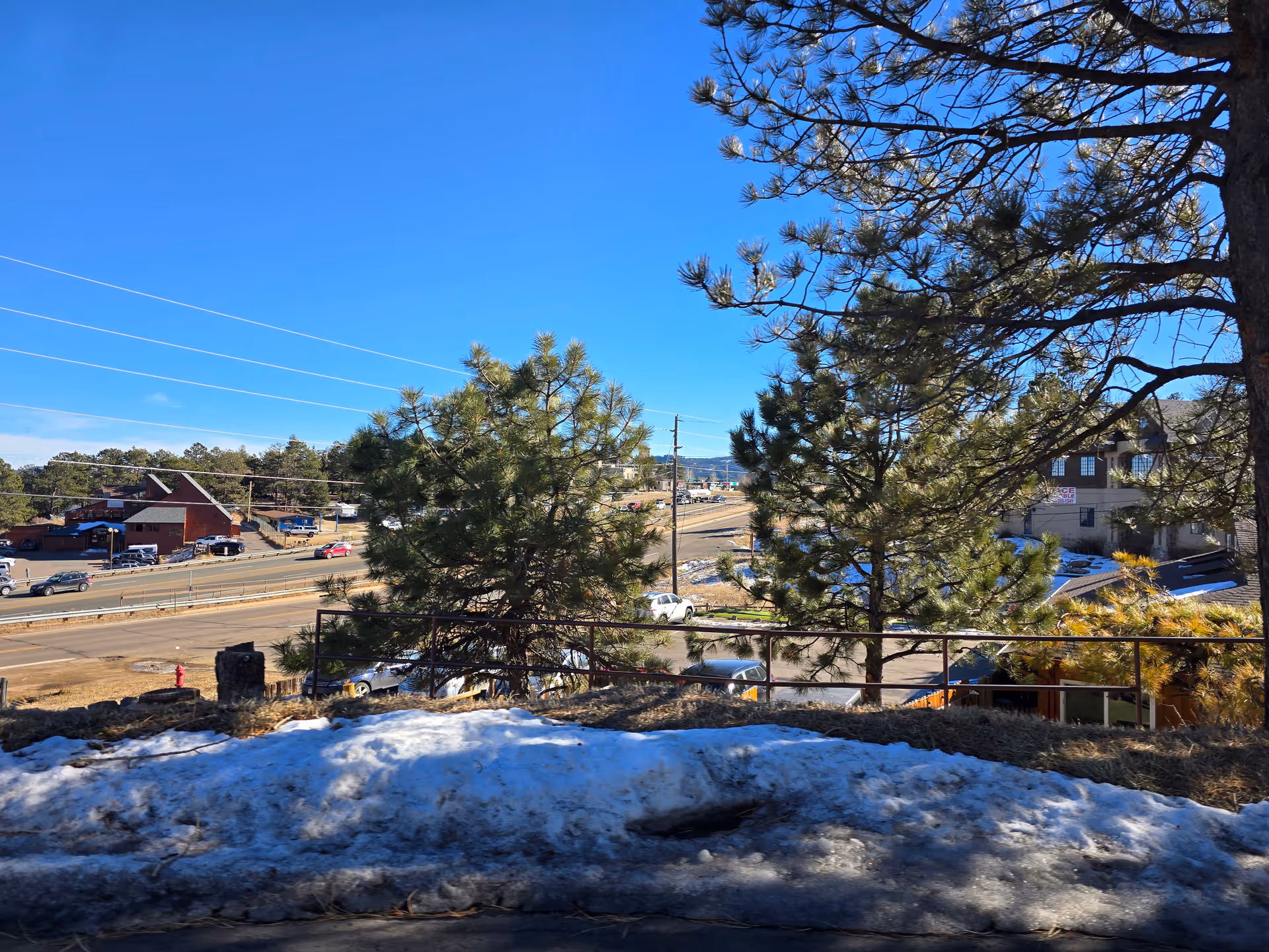 View of a road with several cars driving and parked, surrounded by pine trees and buildings under a clear blue sky. There is a patch of snow on the ground in the foreground.