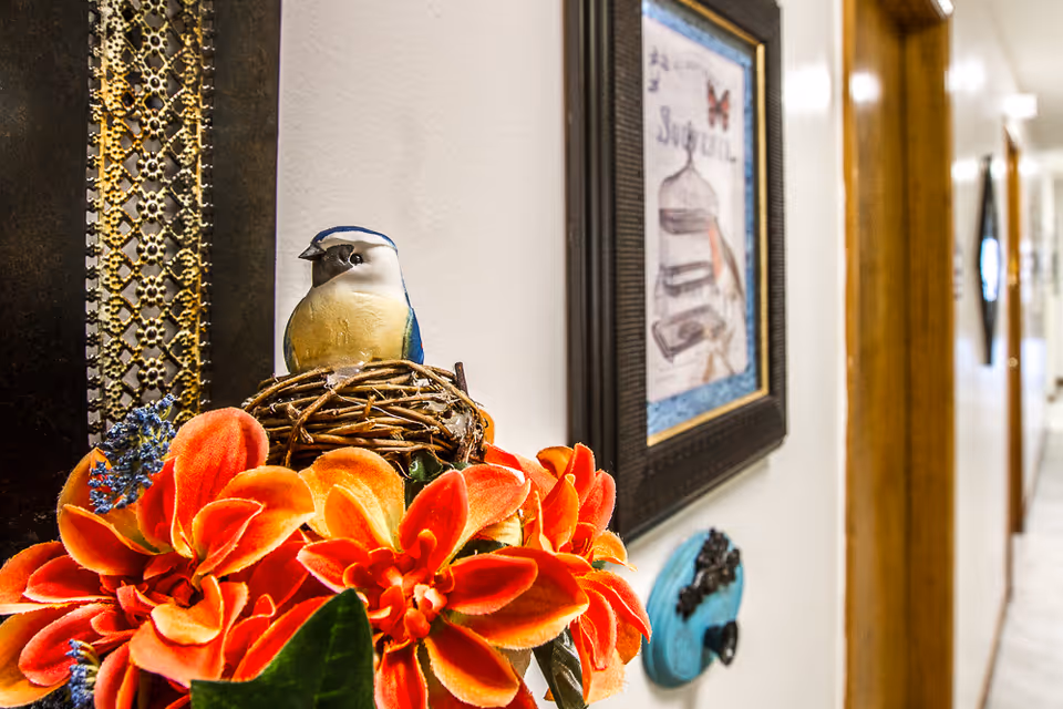 A close-up of a decorative arrangement featuring bright orange flowers and a small bird figurine perched on a nest. In the background, there is a framed picture on a white wall and a hallway with wooden door frames.
