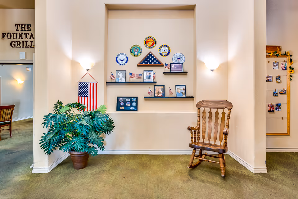 A senior living common area featuring a wall display of military memorabilia and American flags, a wooden rocking chair, and a potted plant.