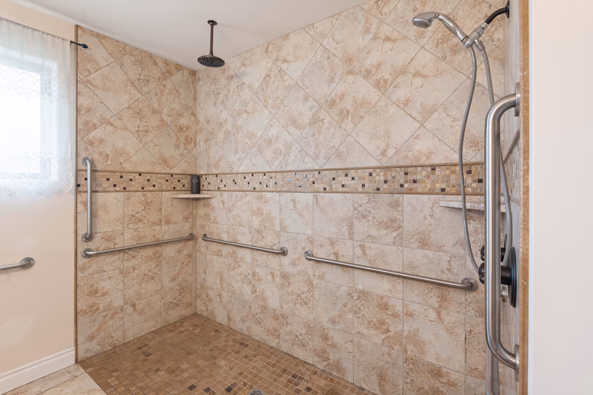 A tiled walk-in shower with beige and brown tiles, featuring multiple stainless steel grab bars on the walls for support. There is a rain showerhead mounted on the ceiling and a handheld showerhead attached to the wall. A small corner shelf holds a dark-colored bottle. A window with a sheer lace curtain is visible on the left side.