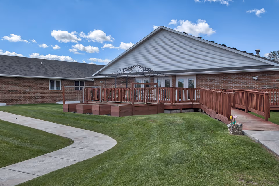 Outdoor view of a senior living facility showing a brick building with a white gabled roof. There is a wooden deck with railings and a ramp leading to the entrance. The deck has a metal frame structure, possibly for a canopy. The surrounding area has well-maintained green grass and a curved concrete walkway. The sky is blue with scattered clouds.