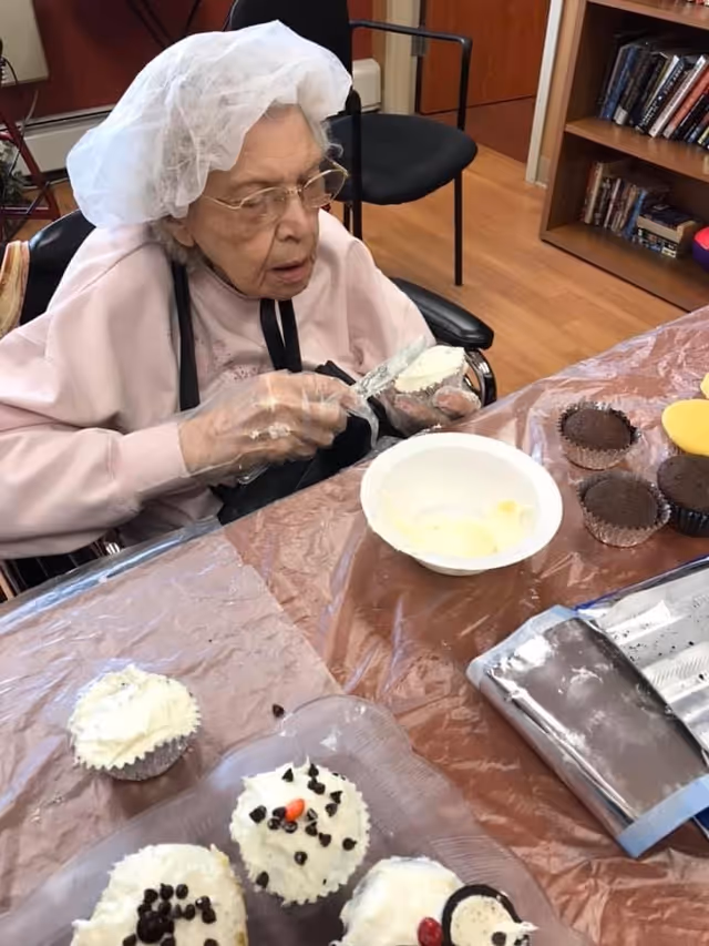 An elderly woman wearing a hairnet and gloves is sitting in a wheelchair at a table, decorating cupcakes with white frosting. Several decorated and undecorated cupcakes are on the table, along with a bowl of frosting and some baking supplies. The setting appears to be an indoor common area with wooden flooring and bookshelves in the background.