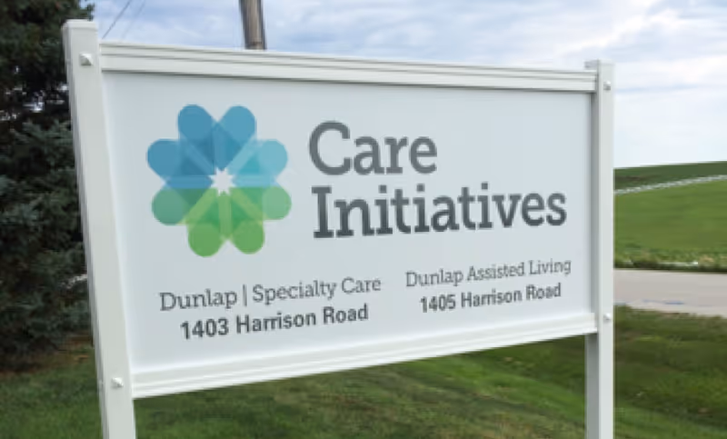 Freestanding outdoor sign reading "Care Initiatives" and listing Dunlap Specialty Care and Dunlap Assisted Living addresses by a grassy roadside.