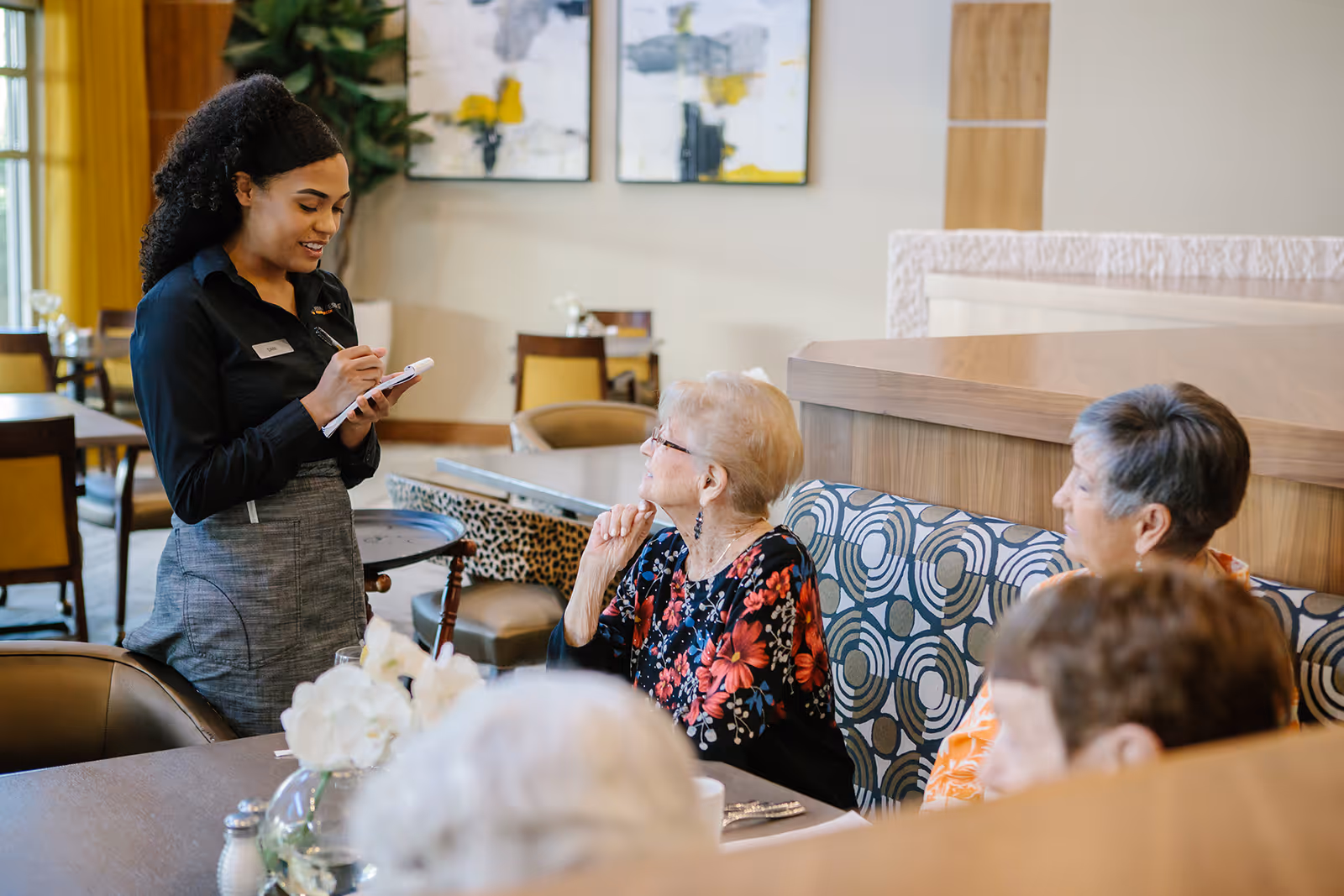 A server takes an order from elderly residents seated at a dining table in a communal dining area.