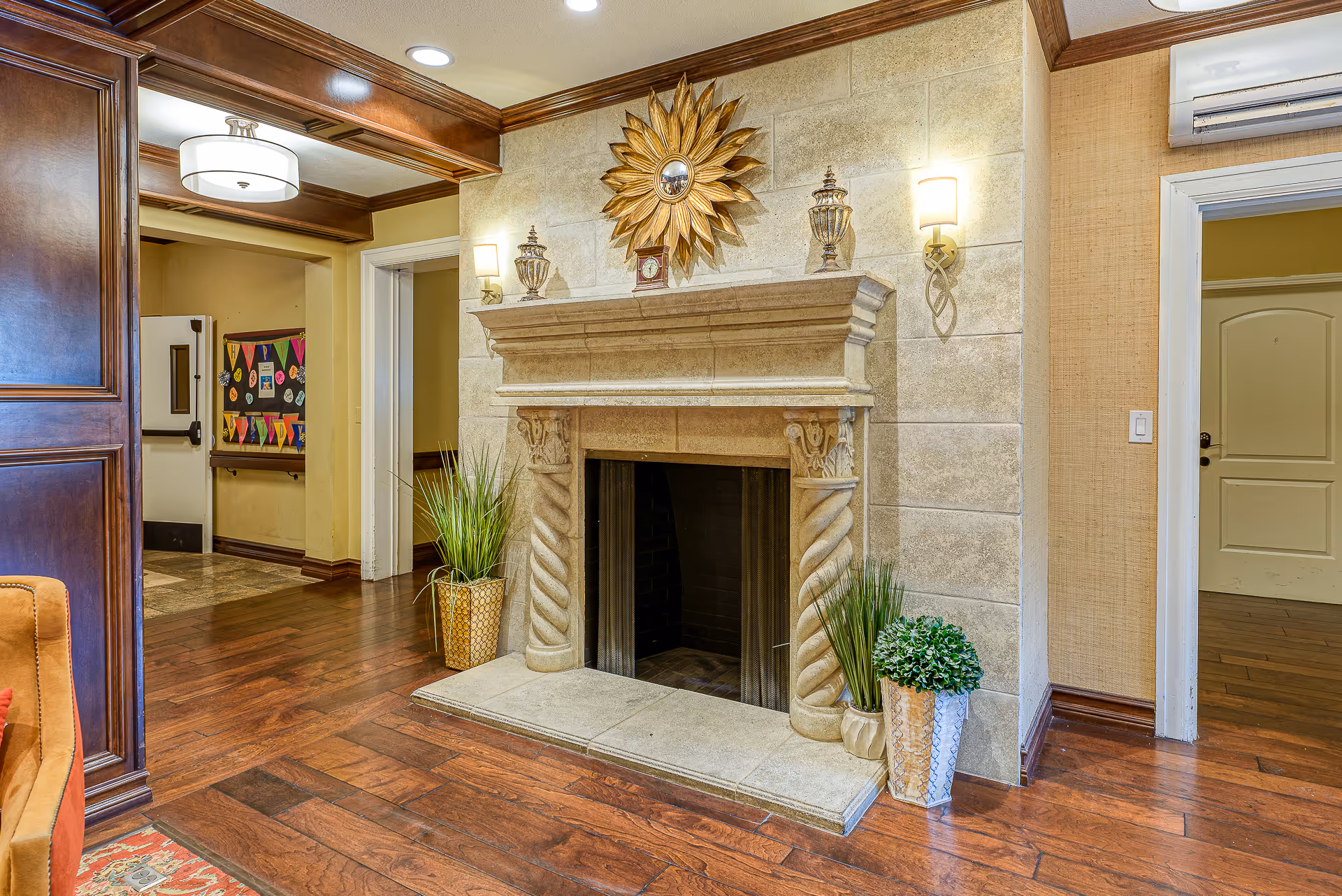 Decorative stone fireplace with twisted columns, potted plants, a sunburst mirror, and hardwood floors in a communal interior room with adjacent doorways.