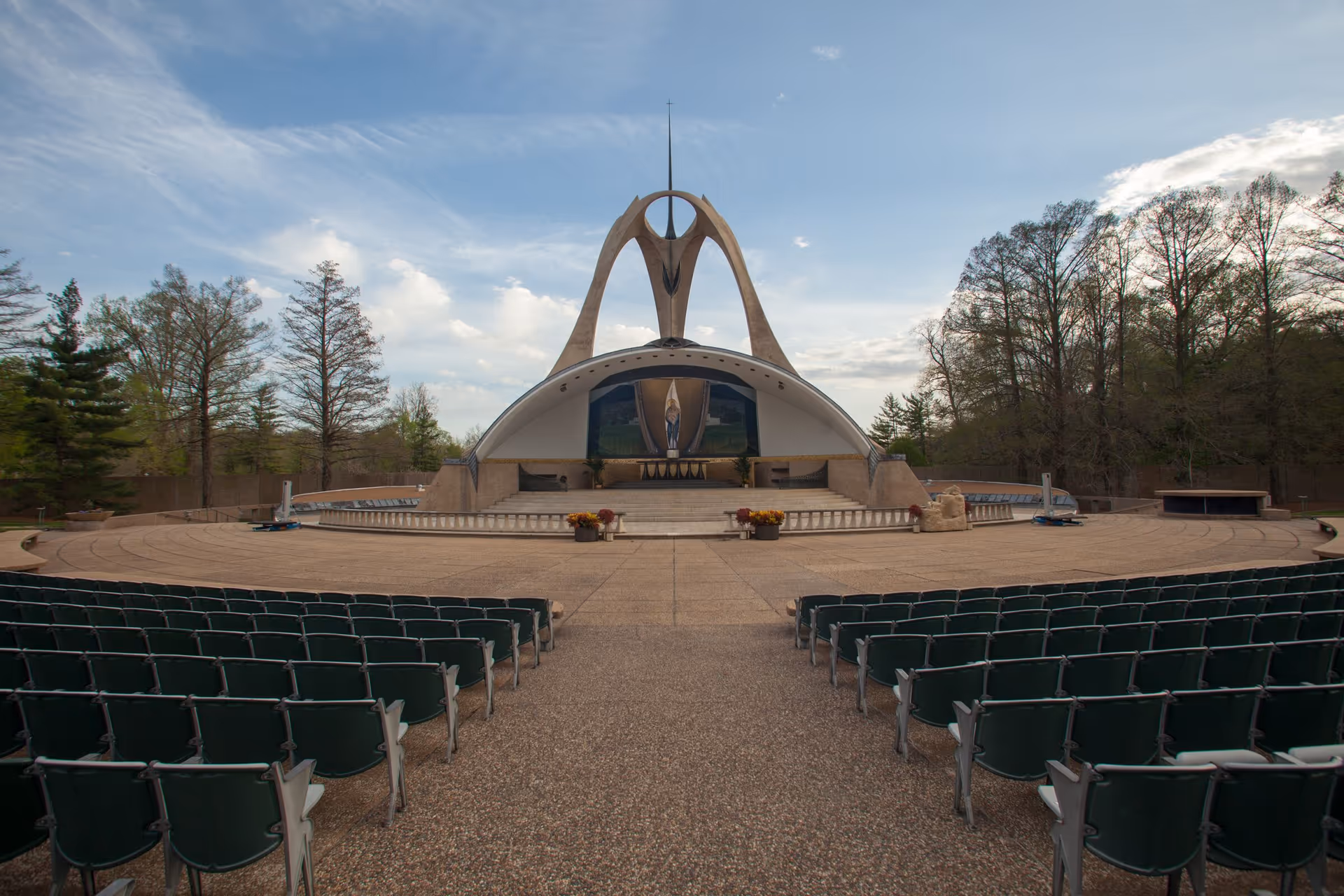 Outdoor amphitheater with rows of green chairs facing a large, modern, curved stage structure with a religious statue and artwork under a partly cloudy sky, surrounded by trees.