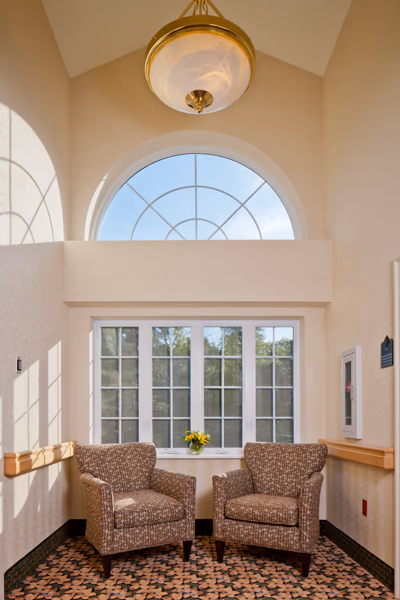 A cozy seating area with two patterned armchairs placed side by side in front of a large window with multiple panes. Above the window is a large arched window letting in natural light. A small vase with yellow flowers sits on the windowsill. The walls are light-colored, and a decorative ceiling light fixture hangs above. The floor has a patterned carpet.
