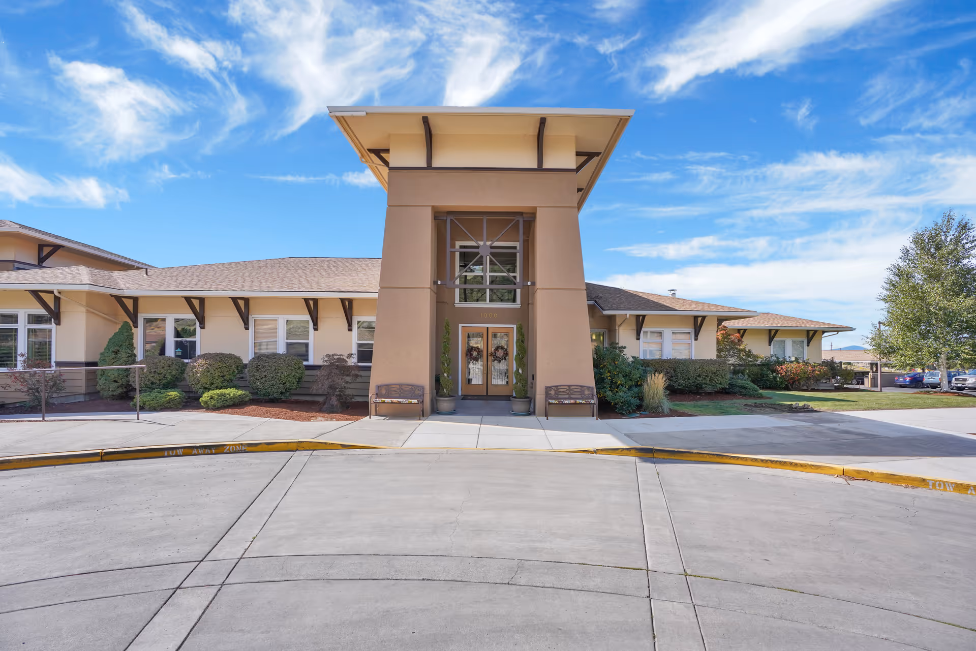 Front exterior view of Crystal Terrace of Klamath Falls, a single-story building with a central entrance featuring double doors and large windows above. The building is surrounded by neatly trimmed bushes and small trees, with a clear blue sky and some clouds in the background. There is a curved driveway and sidewalk leading to the entrance.