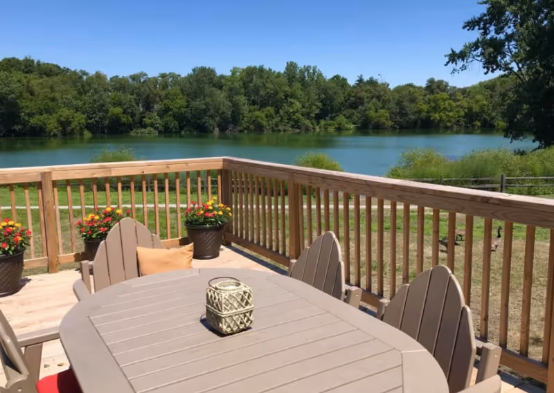 Outdoor wooden deck with a table and chairs, potted flowers, and a scenic view of a lake and trees under a clear blue sky.