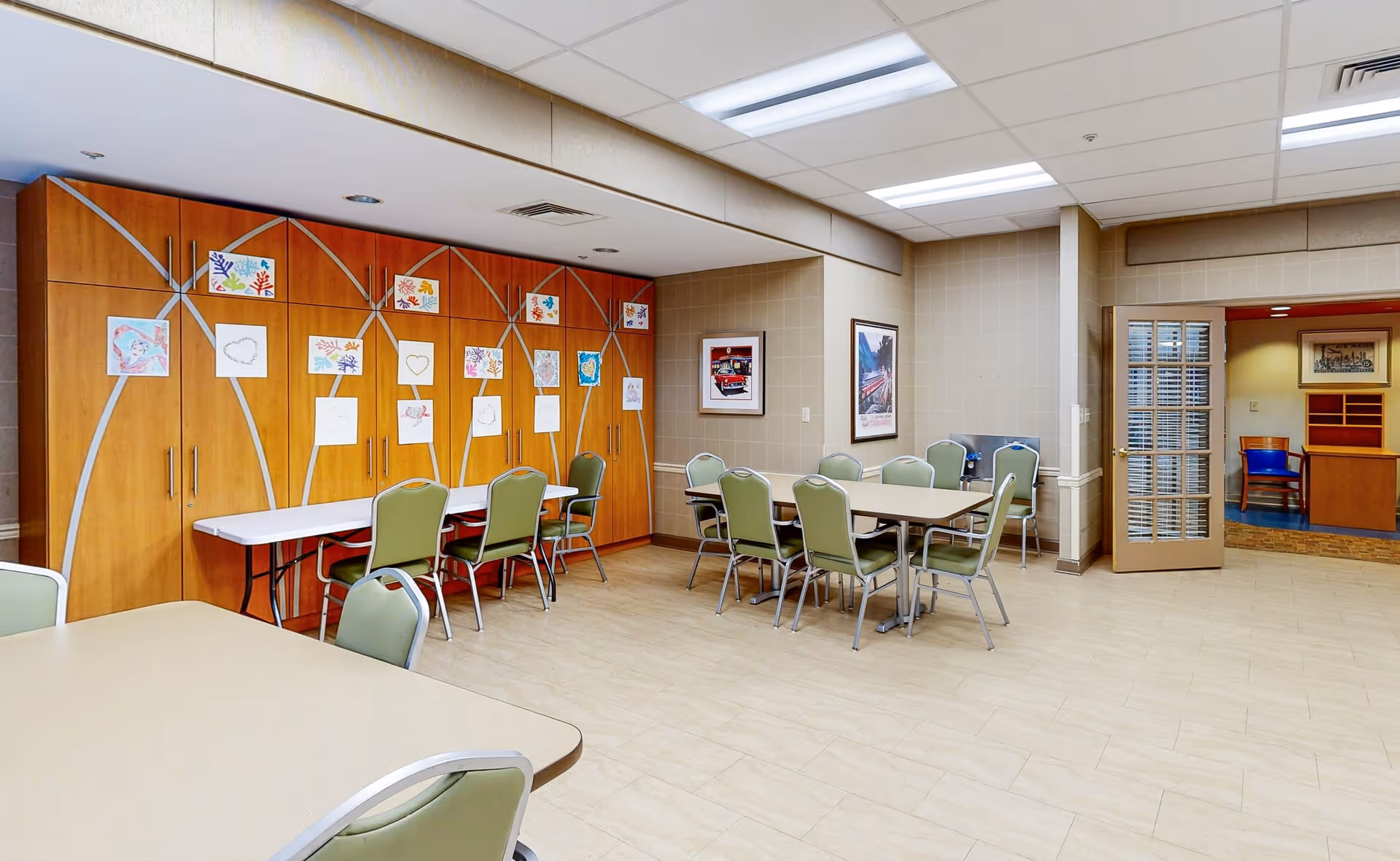 A spacious room with several tables and green chairs arranged around them. The back wall features wooden cabinets decorated with colorful children's drawings and artwork. The room has tiled flooring, beige walls, and fluorescent ceiling lights. There is a door with glass panels leading to another room with blue chairs and wooden furniture.