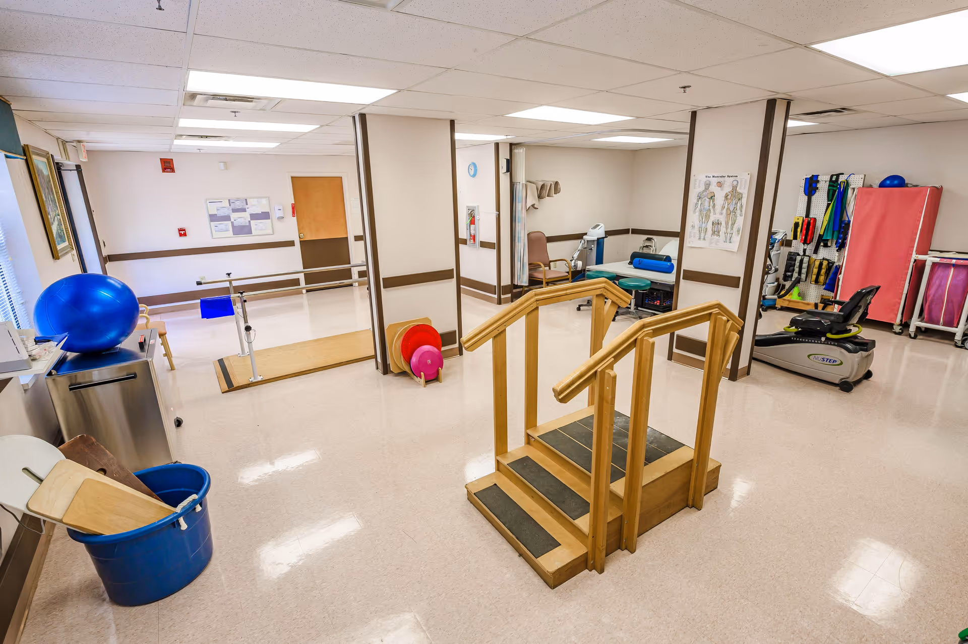 A senior facility physical therapy room with wooden practice stairs, parallel bars, exercise equipment, and therapy balls.