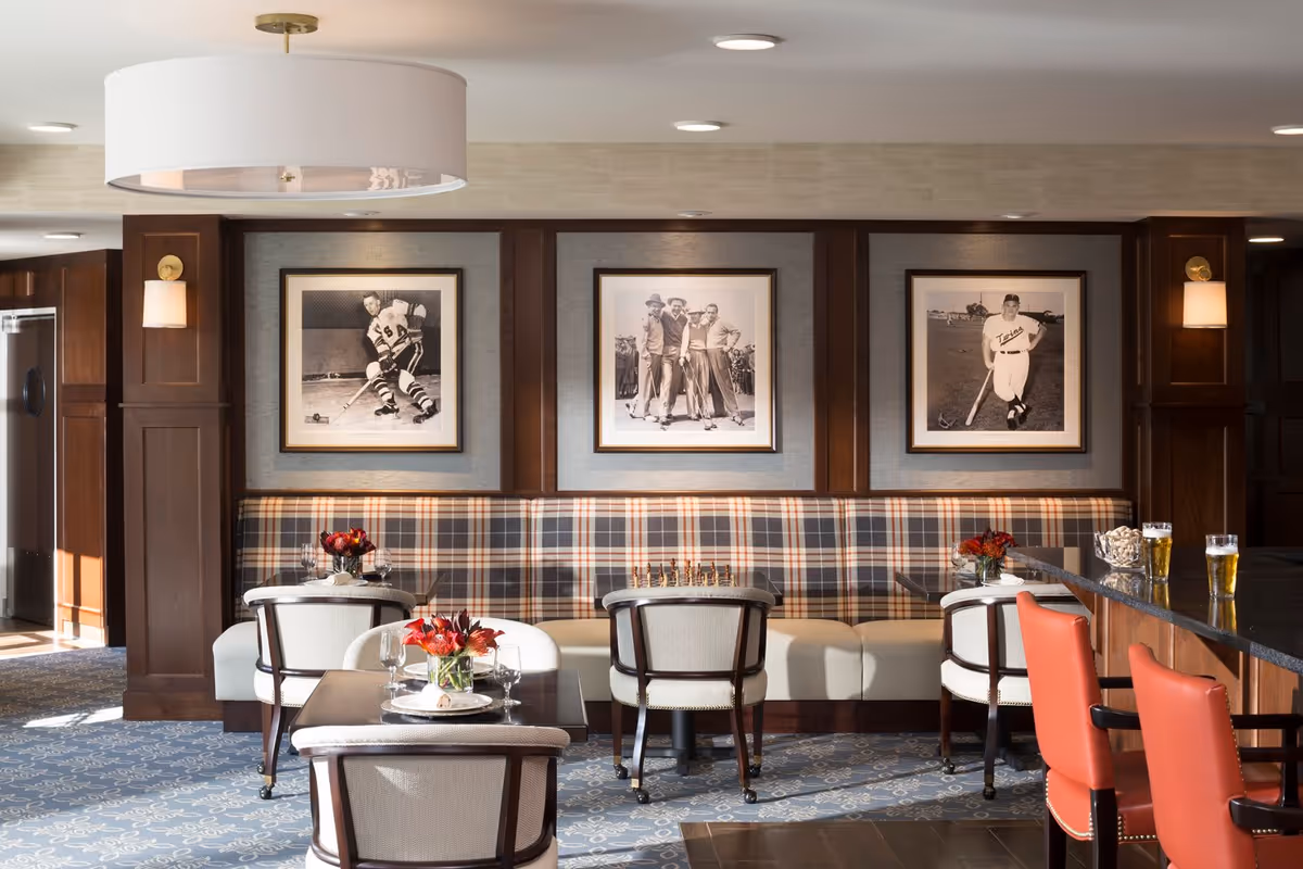 A cozy dining area with a plaid upholstered bench along the wall, three framed black and white sports photographs above it, and several tables with chairs. The tables have small flower arrangements and place settings. To the right, there is a bar counter with red cushioned bar stools and glasses of beer. The room features warm wood paneling and soft lighting.