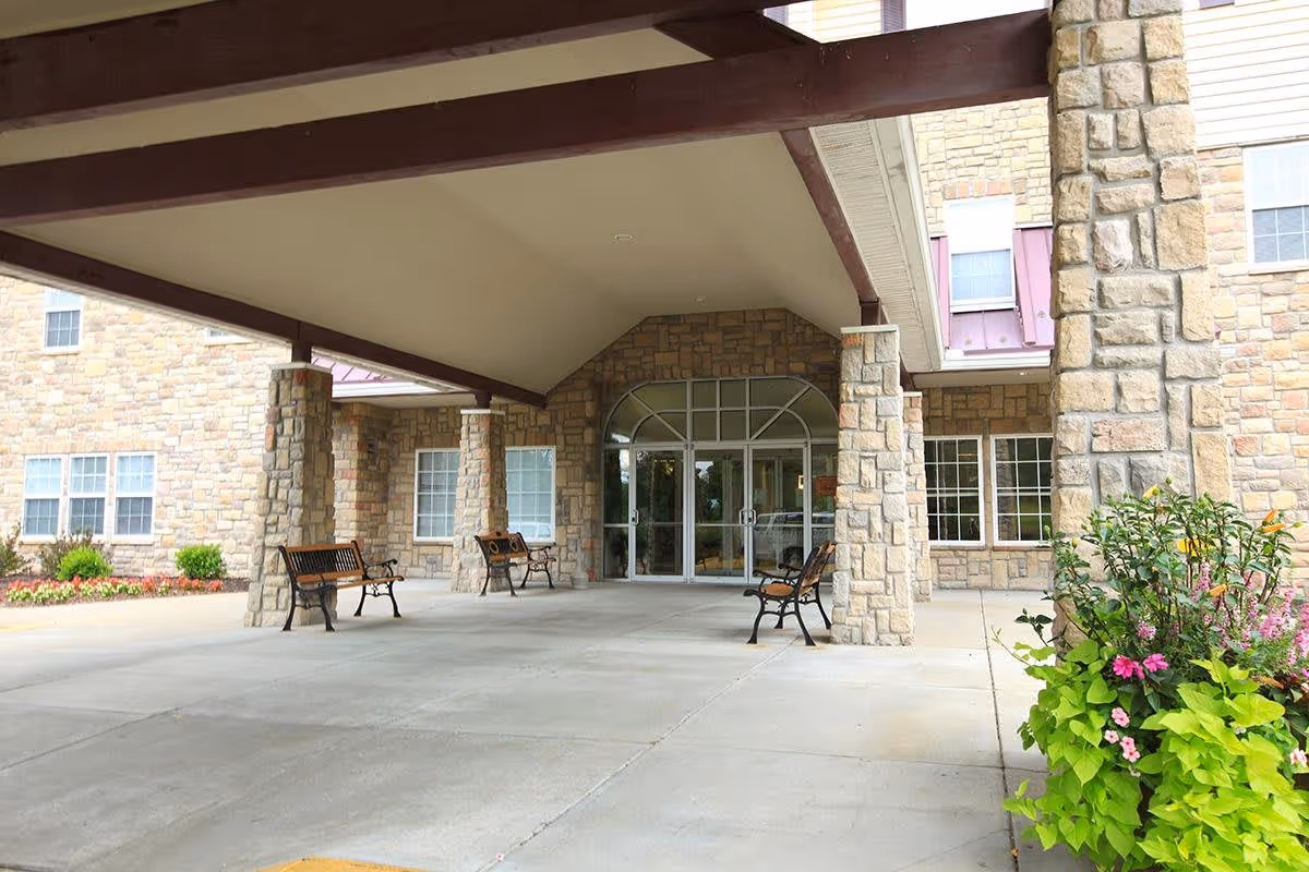 Covered entrance area of a building with stone pillars and walls, glass double doors, several benches, and a flower bed with green and pink plants on the right side.