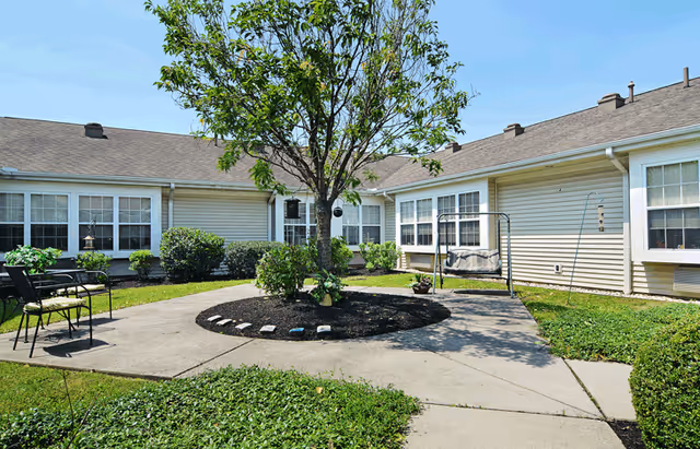 Outdoor courtyard area at Celebration Villa of Lebanon PA featuring a central tree surrounded by a mulched bed, concrete walkways, green grass, bushes, a black metal chair with a cushion, and a cushioned swing bench attached to the building. The building has beige siding and multiple windows.