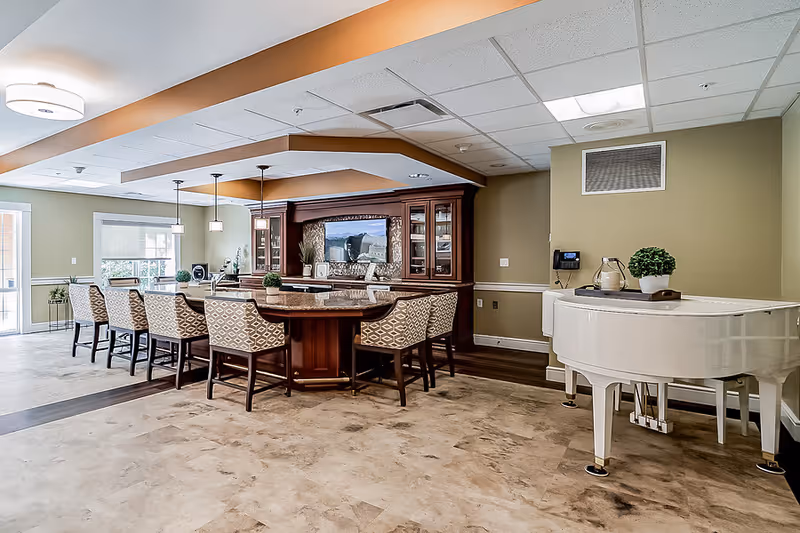 A spacious interior common area featuring a large wooden bar with patterned cushioned chairs arranged around it. The bar has a built-in display cabinet with glass doors and a decorative mirror. To the right, there is a white grand piano with a small plant and decorative items on top. The room has beige walls, a tiled floor, and ceiling lights, with windows allowing natural light to enter.