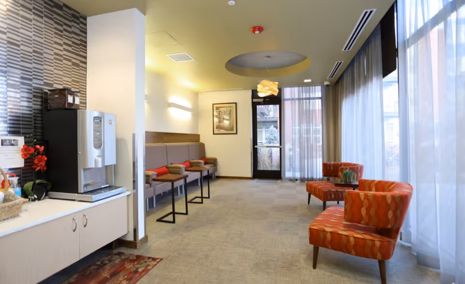 A bright lounge with patterned armchairs, bench seating, a coffee station, and floor-to-ceiling windows.