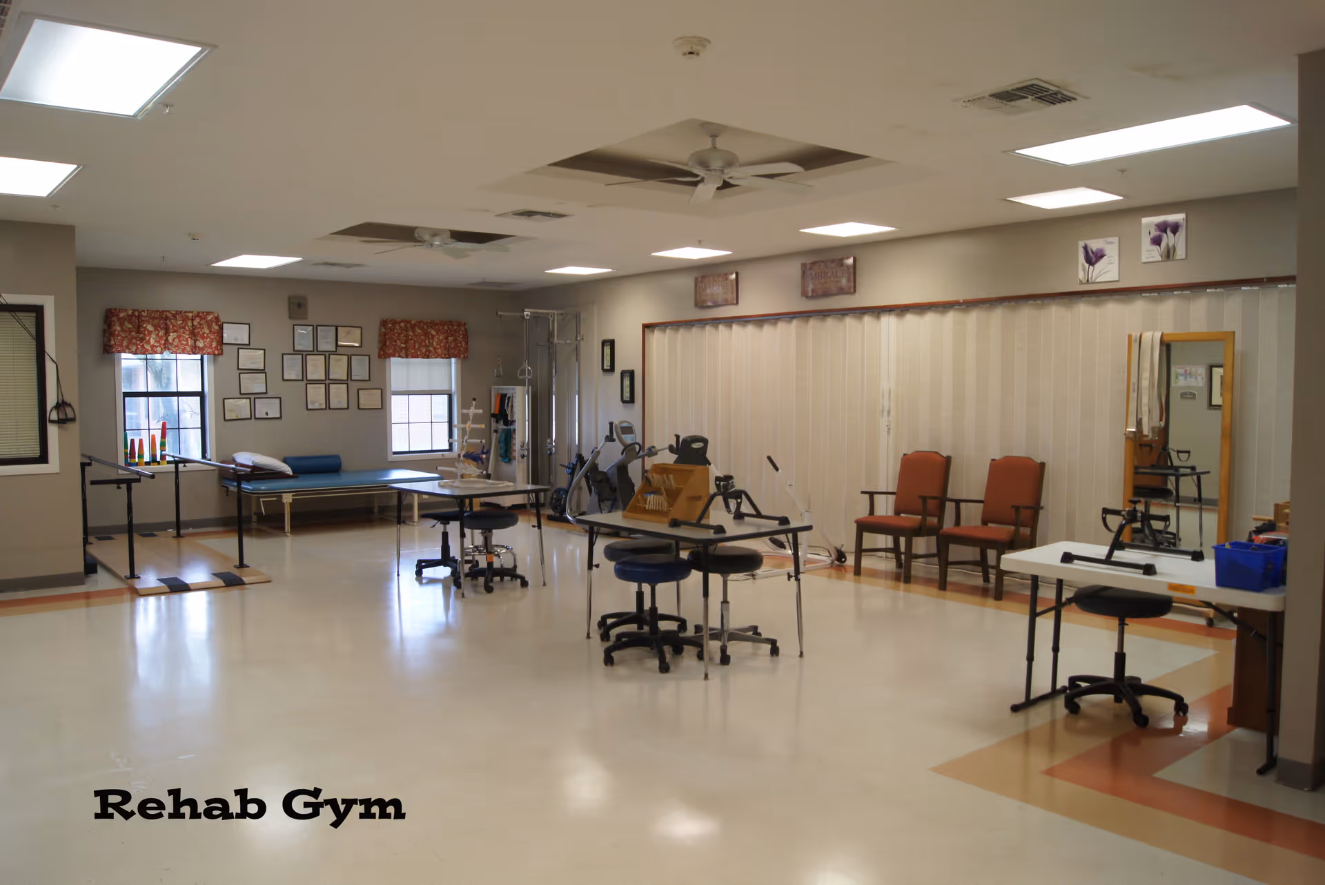 A spacious rehab gym room with exercise equipment including parallel bars, therapy tables, chairs, and various rehabilitation tools. The room has large windows with floral curtains, framed certificates on the wall, ceiling fans, and bright overhead lighting. The floor is clean and has a pattern of beige and orange tiles.