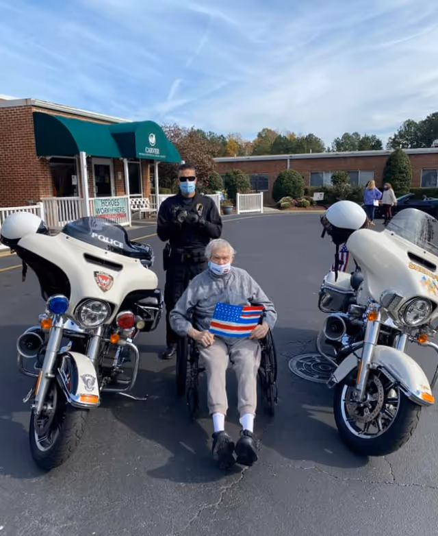 An elderly man in a wheelchair holding an American flag sits between two police motorcycles outside the Carver Rehabilitation & Living Center entrance with a masked officer standing behind him.