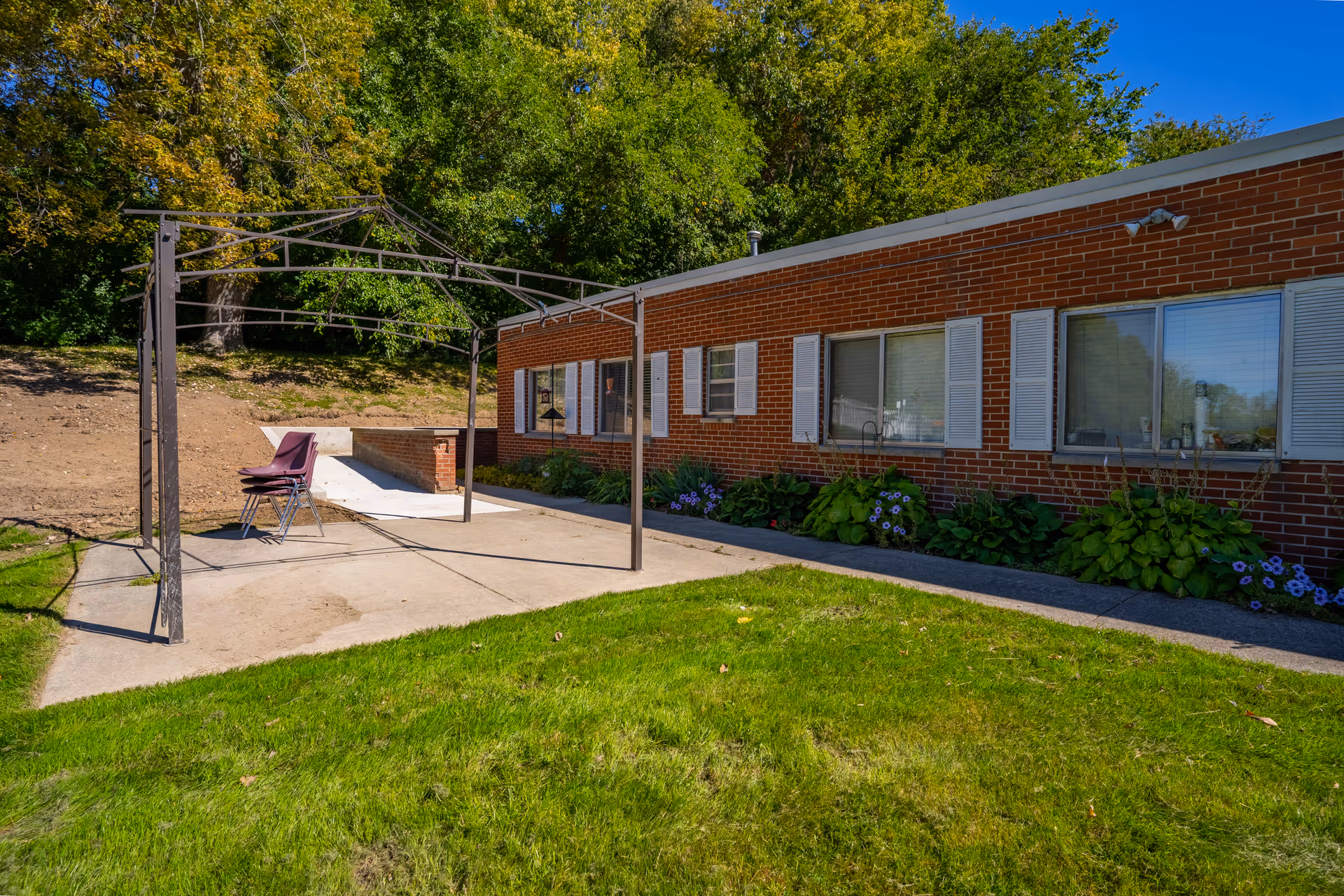 Outdoor area of a brick building with white window shutters, a green lawn, and a metal frame structure with stacked chairs underneath. Trees and a dirt slope are visible in the background.