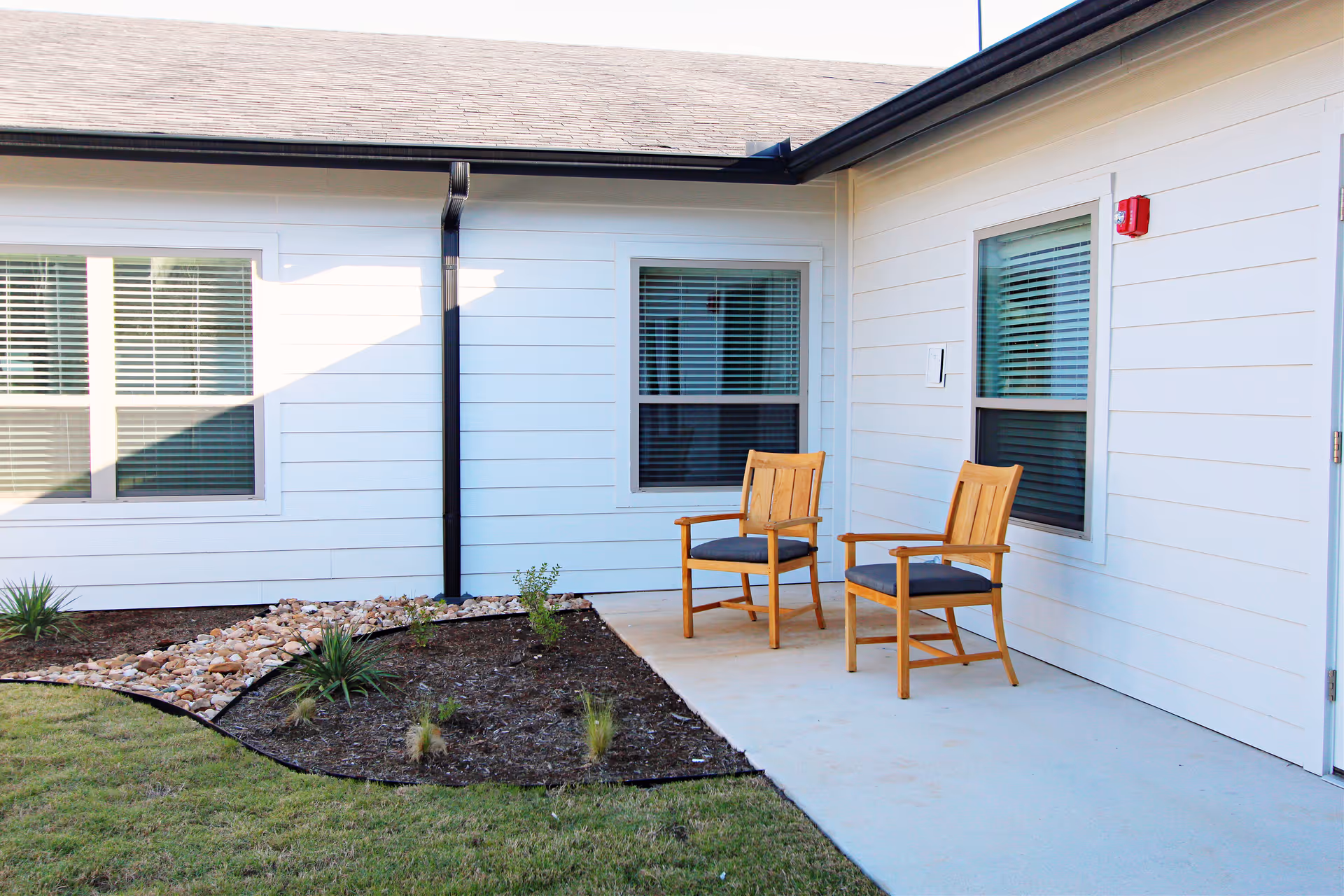 Outdoor patio area at The Haven at San Gabriel with two wooden chairs with cushions placed on a concrete surface next to a white building with windows and a small landscaped garden with plants and rocks.