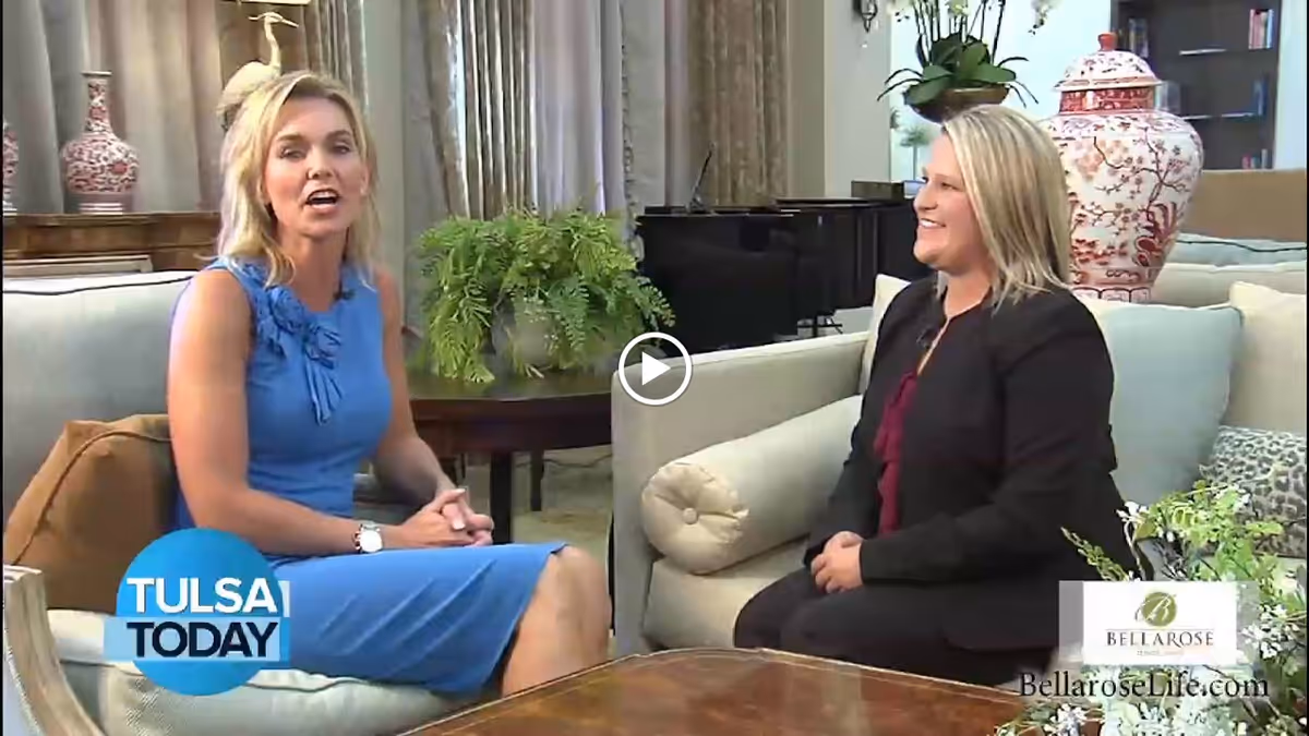 Two women sitting and talking in a well-decorated living room area with sofas, plants, and decorative vases. One woman is wearing a blue dress and the other is in a black jacket. There is a piano in the background.