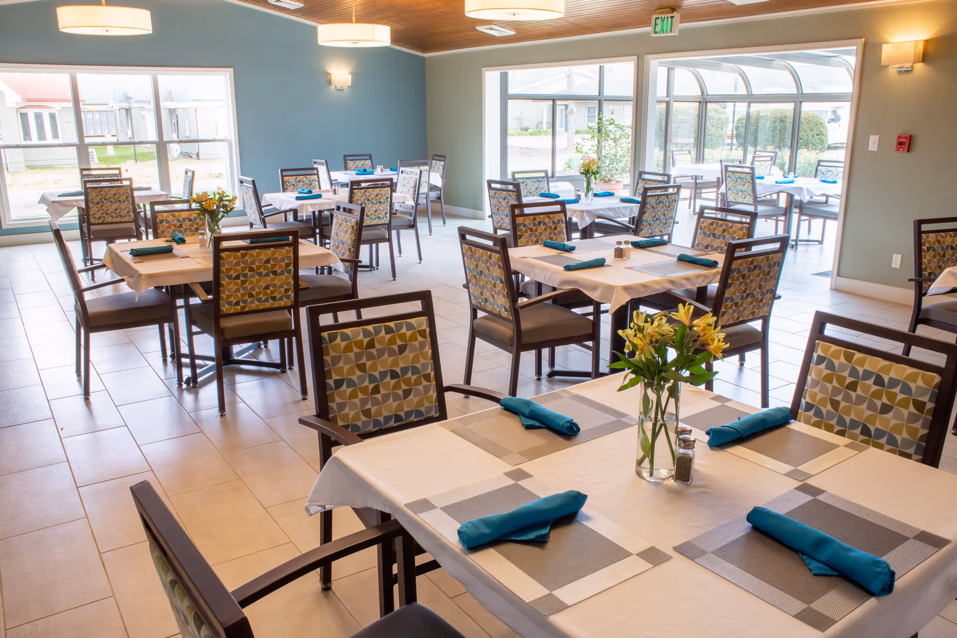 A bright dining room with multiple tables covered with white tablecloths and gray placemats, each set with teal napkins and a small vase of yellow flowers. The room has large windows letting in natural light and a glass door leading to an outdoor area. The chairs have patterned upholstery with geometric designs.