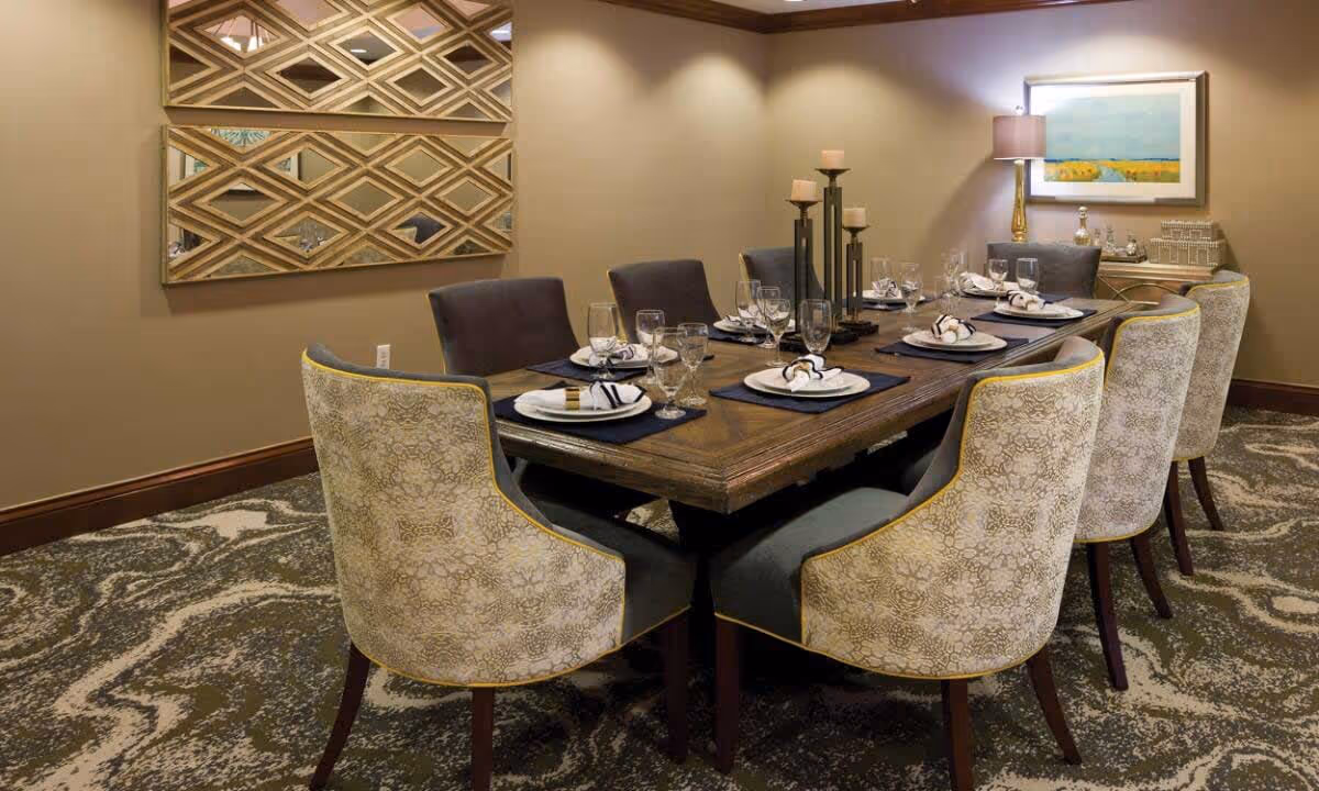 A formal dining room with a long wooden table set for eight people. The table is arranged with plates, napkins, glasses, and candle holders. The chairs around the table have patterned upholstery with a yellow trim. The room has a patterned carpet, beige walls, a decorative mirror on one wall, and a framed painting with a lamp on a side table.