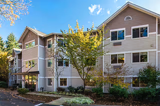 Three-story assisted living building with beige siding and surrounding trees.