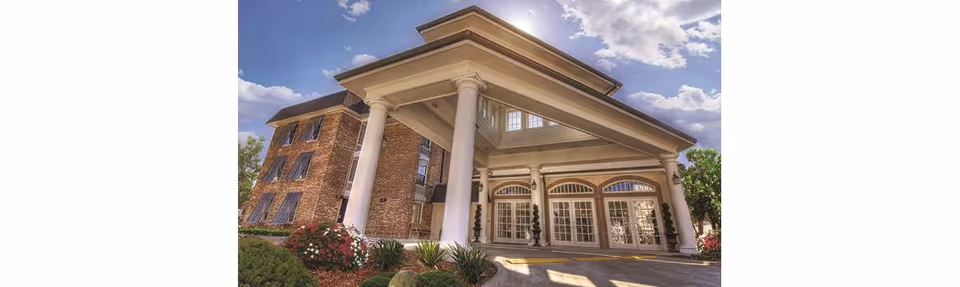Exterior view of a large brick building with tall white columns supporting a covered entrance. The building has multiple windows with awnings and is surrounded by landscaped greenery and flowers under a partly cloudy sky.