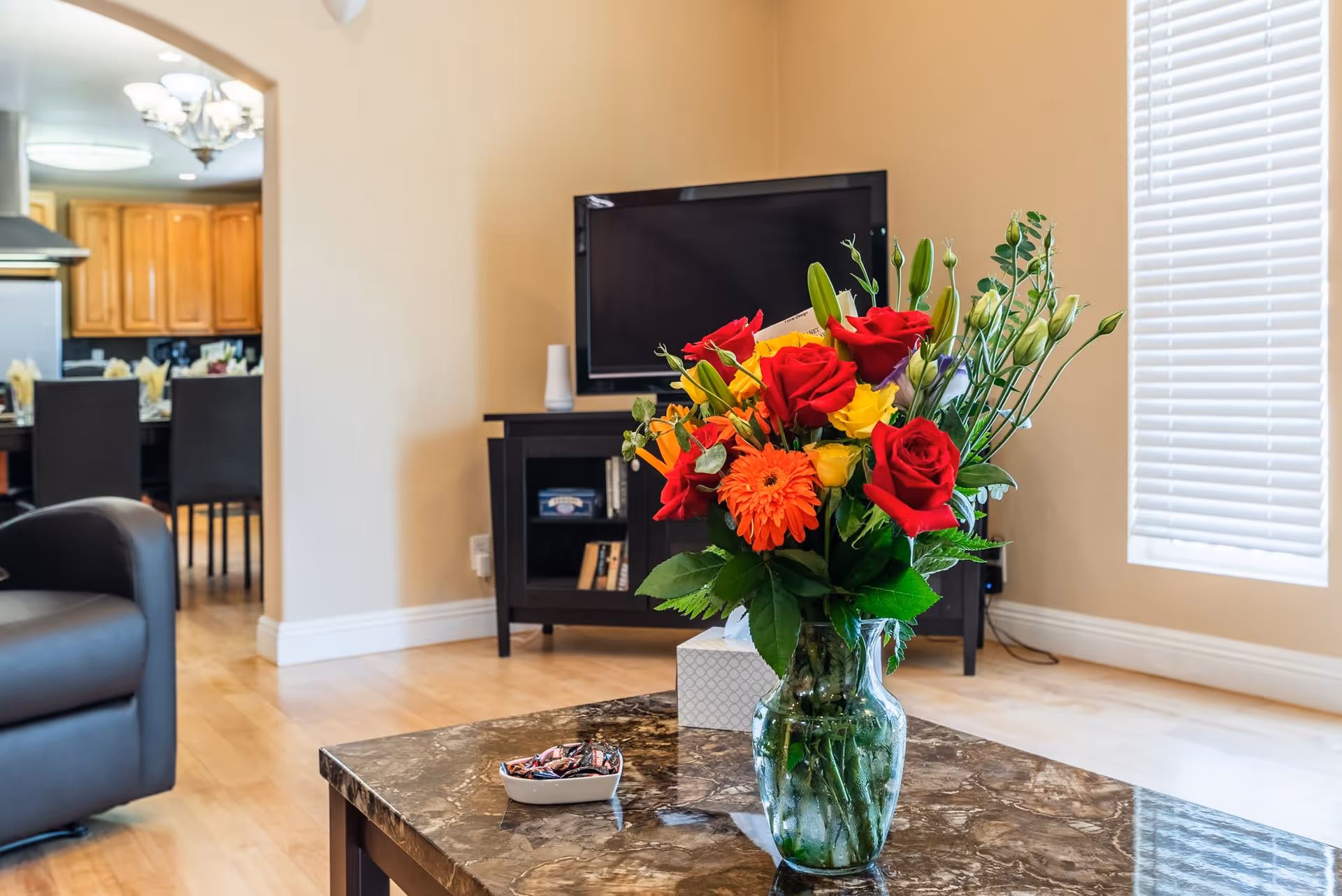 A living room area with a marble coffee table holding a vase of colorful flowers including red roses and orange gerbera daisies. In the background, there is a black TV stand with a flat-screen TV and some books. To the left, a black leather chair is partially visible, and through an open doorway, a dining area with a table and chairs is seen. The room has light wood flooring and beige walls with a window covered by white blinds.