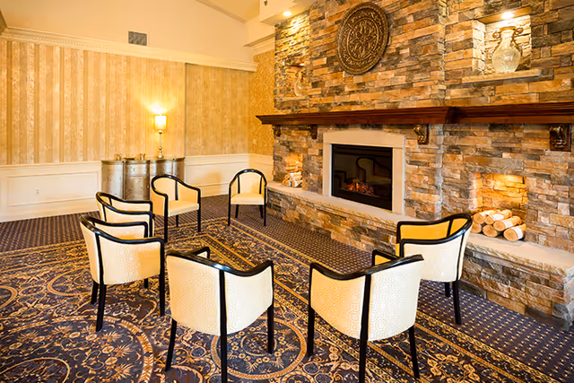 A cozy seating area with six cream-colored chairs arranged in a circle on a patterned carpet. The room features a stone fireplace with a wooden mantel, decorative wall sconces, and a round ornamental wall piece above the fireplace. The walls have a warm beige wallpaper and there is a small table with a lamp in the corner.