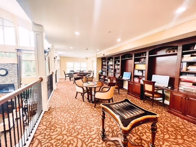 A spacious, well-lit common area with patterned carpet, multiple round tables with chairs, built-in wooden bookshelves, and computer workstations. The room overlooks a lower level with a stone fireplace and large windows letting in natural light.