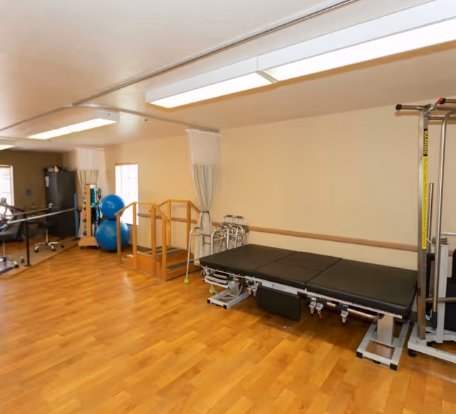 A rehabilitation therapy room with wooden flooring, a black adjustable therapy table, parallel bars for walking exercises, blue exercise balls, walkers, and a metal exercise machine. The room has beige walls and fluorescent ceiling lights.