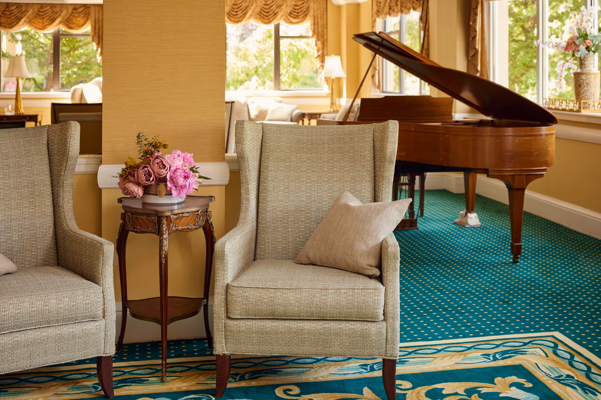 A cozy interior lounge area featuring two beige upholstered armchairs with a small ornate wooden side table between them holding a vase of pink and purple flowers. In the background, there is a grand piano near large windows with yellow curtains, and a patterned green carpet covers the floor.
