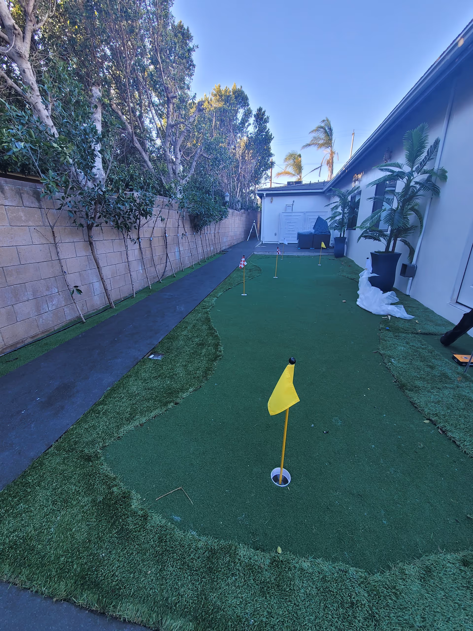 Outdoor putting green area with several small golf holes marked by yellow and checkered flags, surrounded by artificial grass. There is a paved walkway on the left side and a beige brick wall with trees behind it. On the right side, there is a building wall with potted plants and some outdoor furniture in the background under a clear blue sky.
