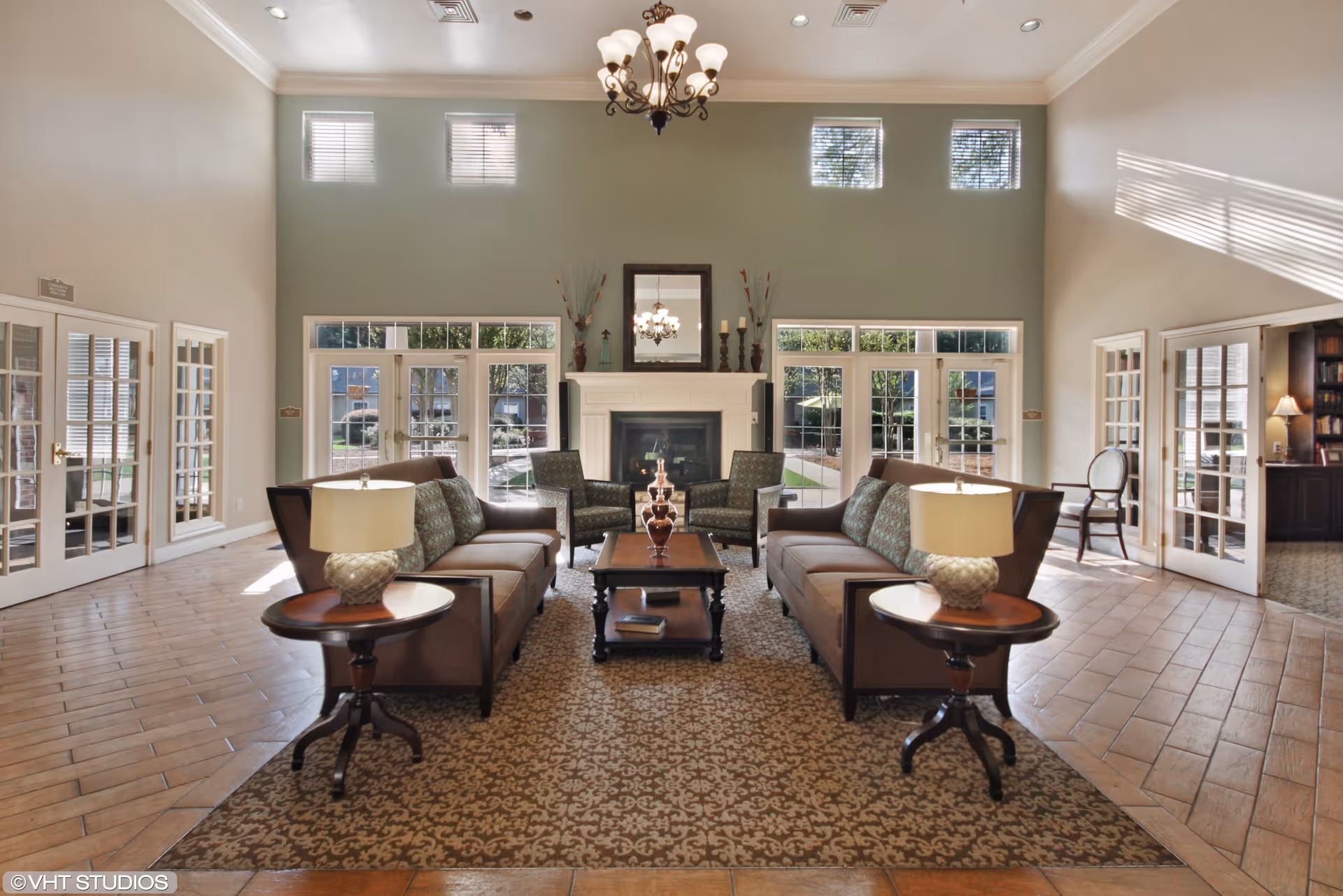 A spacious and well-lit living room area with two brown sofas facing each other, two armchairs, a coffee table in the center, and side tables with lamps. The room features large windows and glass doors letting in natural light, a fireplace with decorative items on the mantel, and a chandelier hanging from the ceiling.