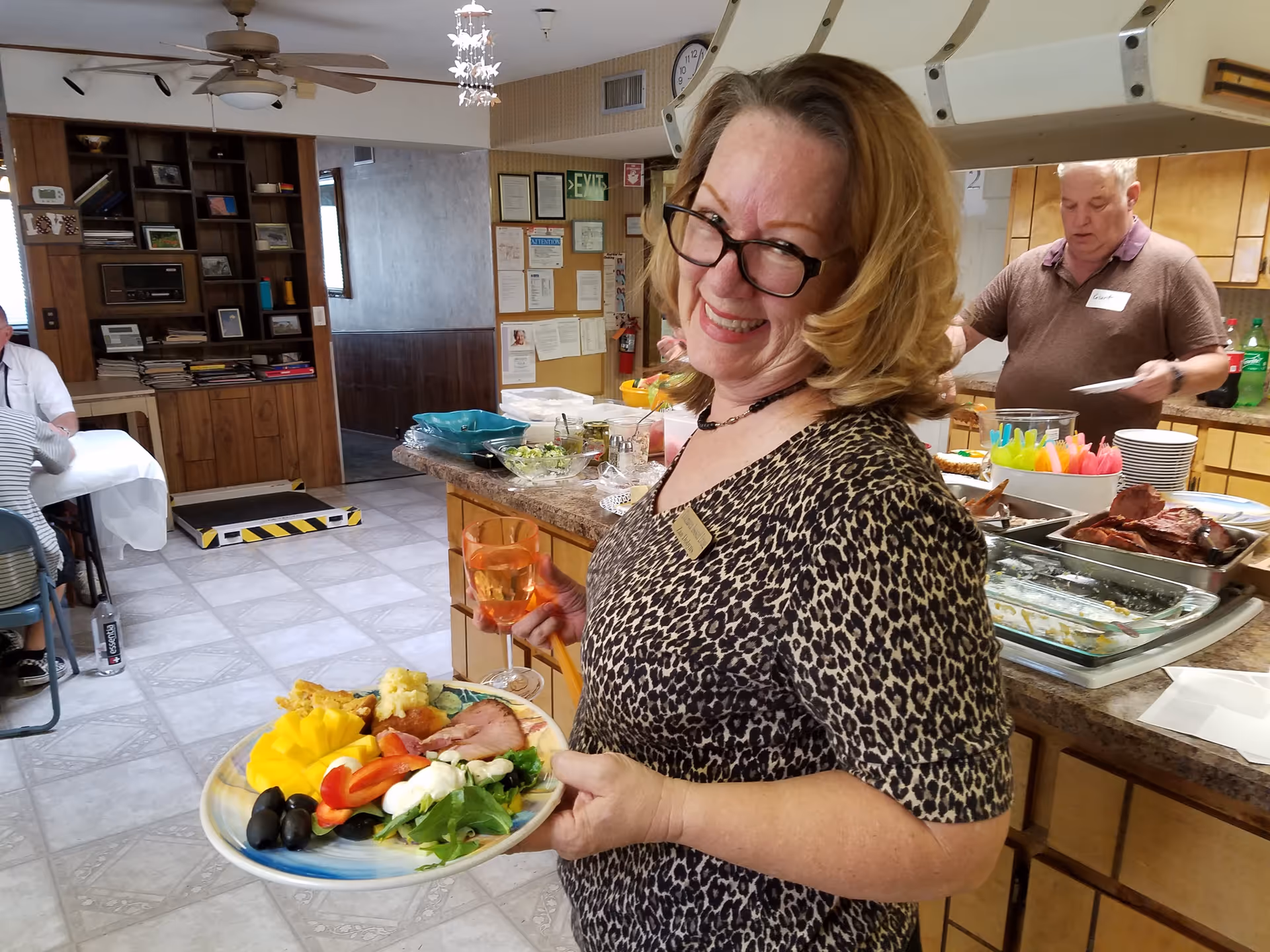 A smiling woman holding a plate of food and a drink stands in front of a buffet in a communal dining area.
