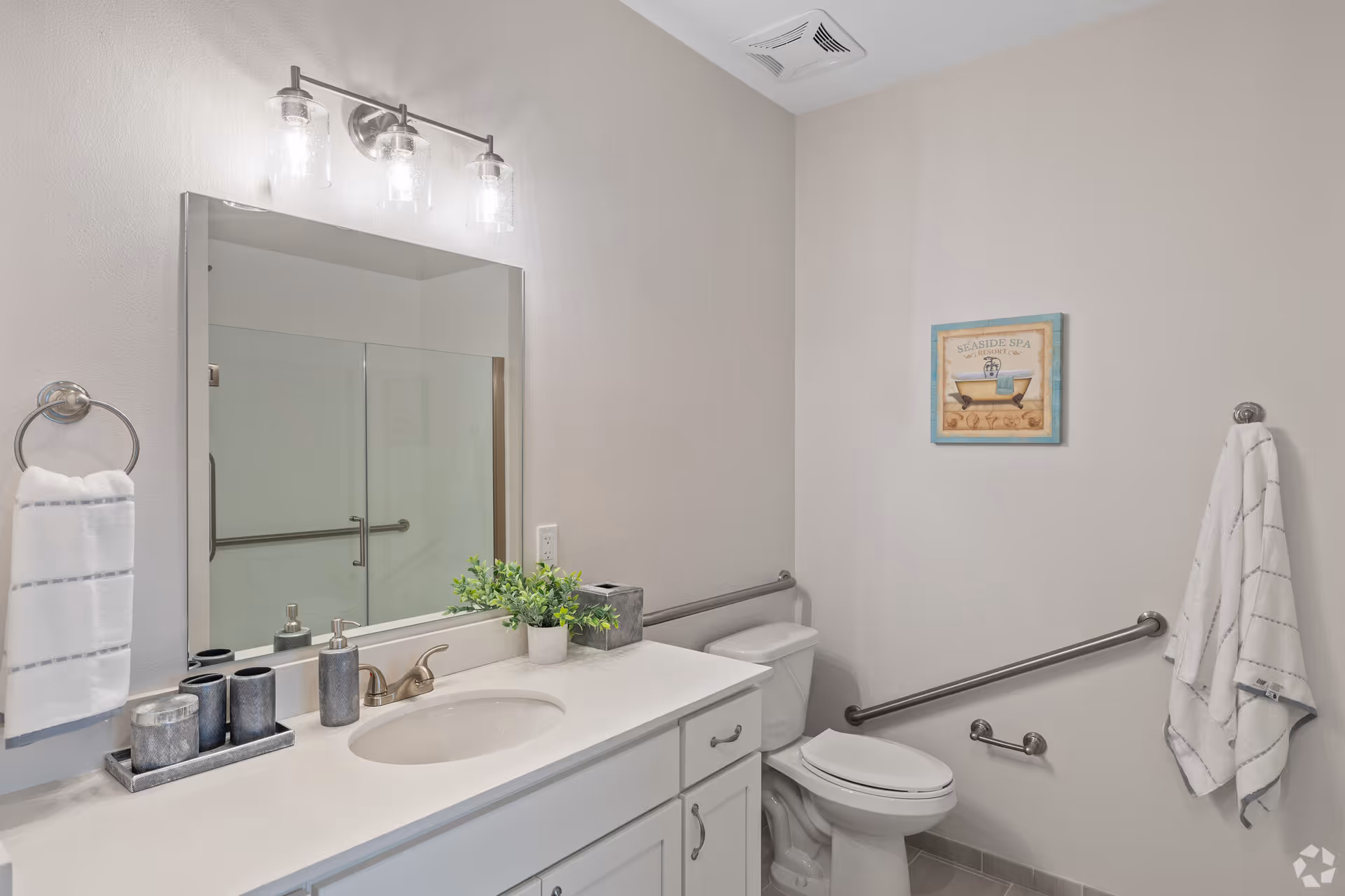 A clean and modern bathroom featuring a white vanity with a sink, a large mirror with three light fixtures above it, a toilet with grab bars on the wall, a towel ring with a white towel, a wall-mounted towel hook with a white towel, a small potted plant, and a framed seaside spa picture on the wall.