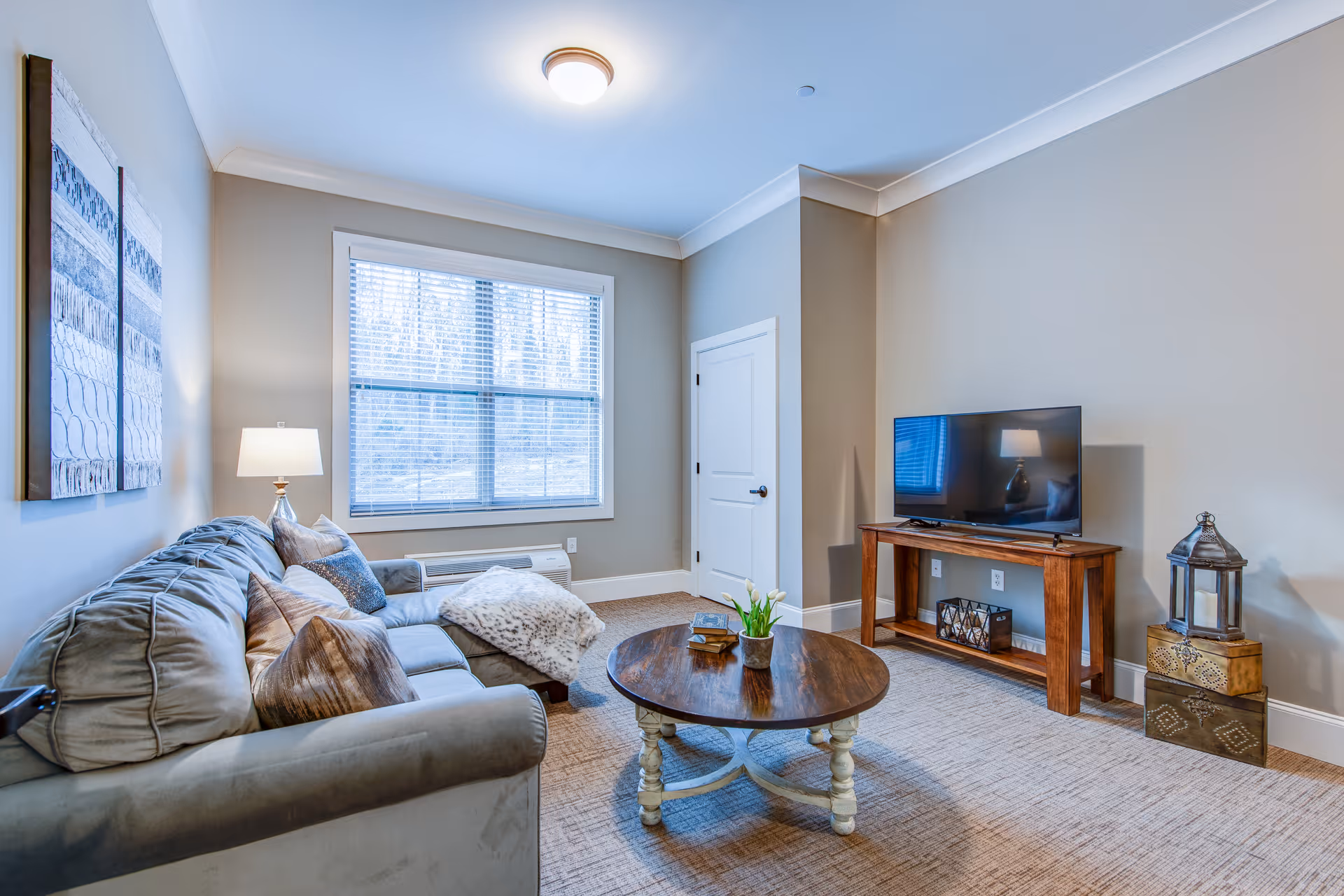 A cozy living room with a gray sofa adorned with multiple pillows and a throw blanket. A round wooden coffee table with a small plant and decorative items sits in front of the sofa. Across from the sofa is a wooden TV stand with a flat-screen television and decorative lanterns on the floor beside it. The room has beige walls, a window with blinds, a ceiling light, and a closed white door.