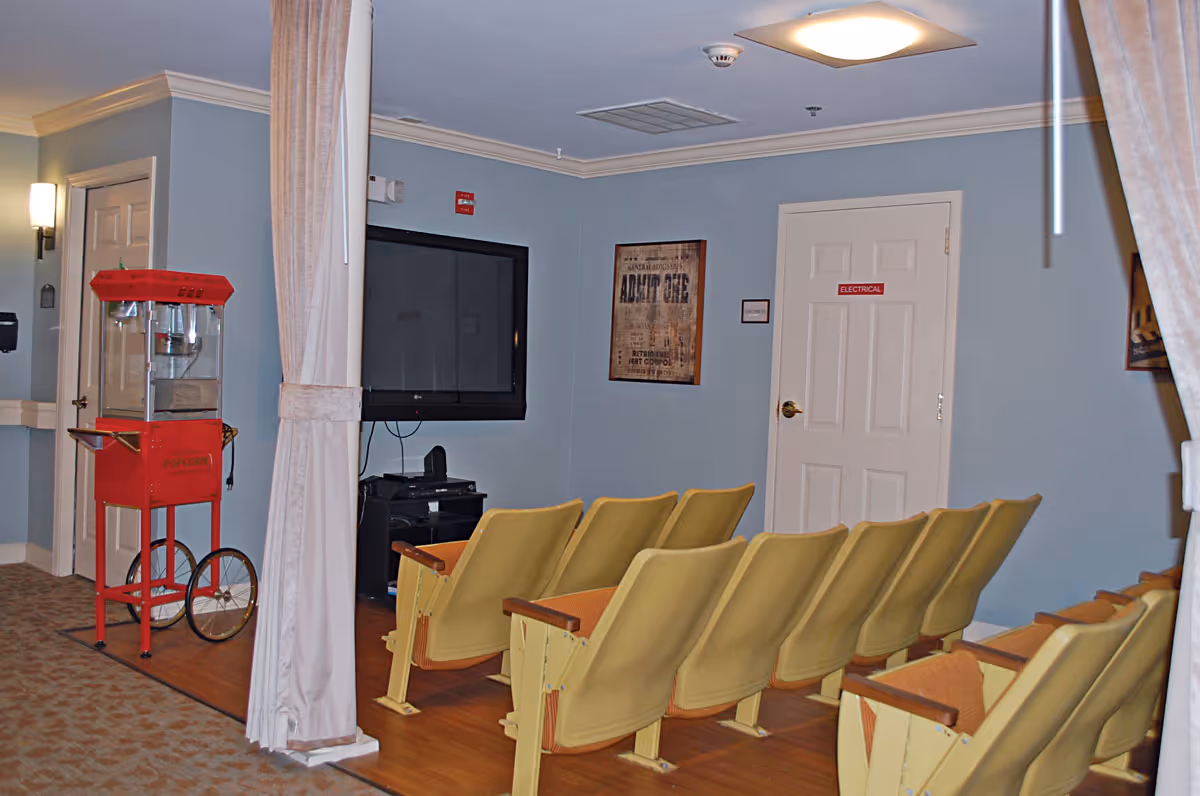 A small theater room with two rows of yellow theater-style seats facing a large flat-screen TV mounted on a light blue wall. There is a red popcorn machine on wheels to the left side of the room. The room has light blue walls, a white door labeled 'ELECTRICAL,' and framed posters on the walls. Beige curtains partially frame the room.