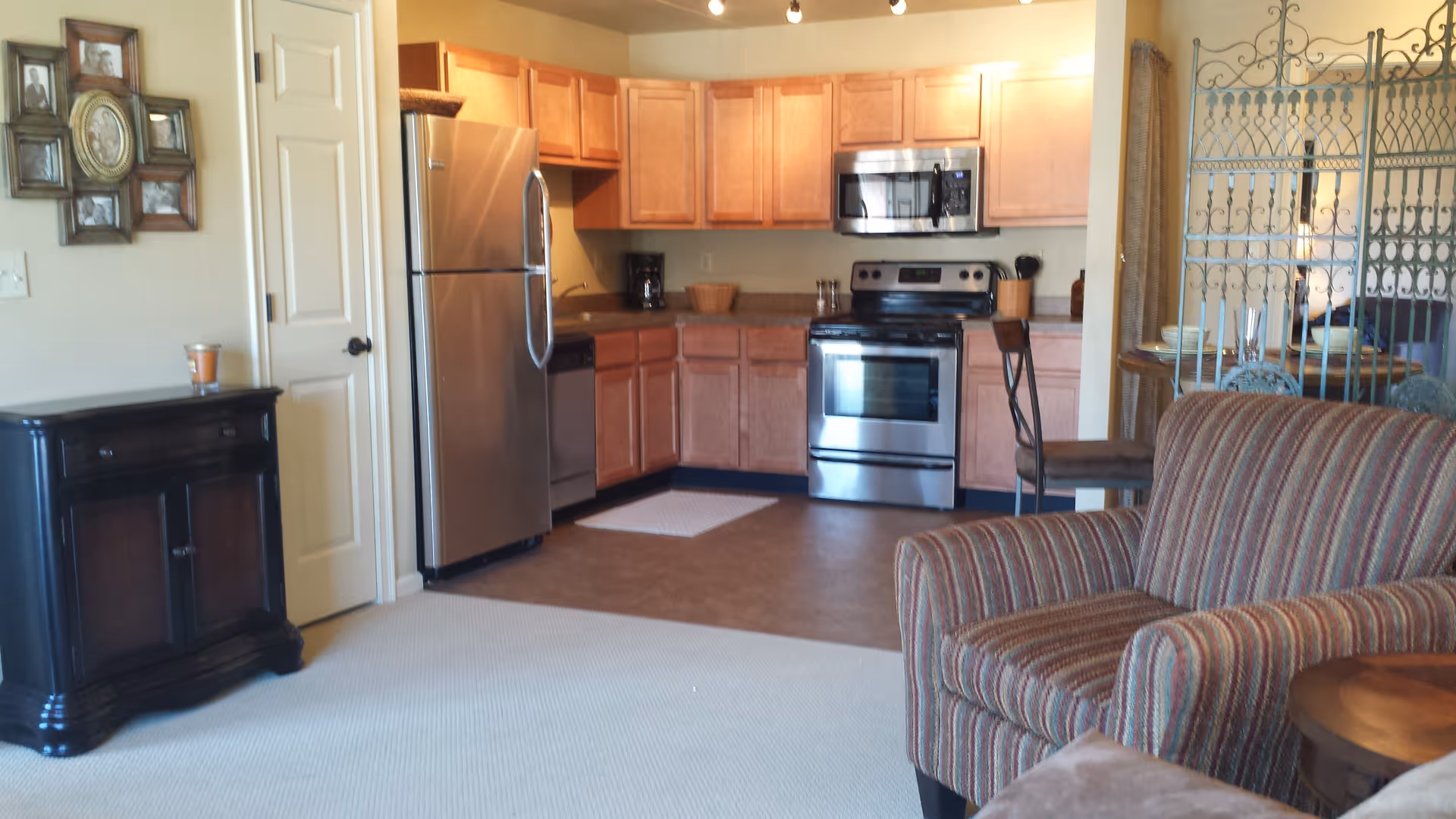Interior view of a senior living facility showing a kitchen area with wooden cabinets, stainless steel refrigerator, oven, and microwave. Adjacent to the kitchen is a living area with a striped armchair and a decorative metal room divider. A small dark wooden cabinet with a candle on top is against the wall, and a photo frame collage hangs above it.