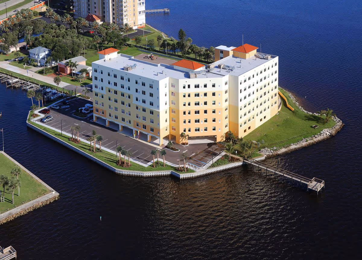 Aerial view of a multi-story senior living facility named Grand Villa of Palm Bay, located on a waterfront with a dock extending into the water. The building is surrounded by parking spaces, palm trees, and green lawns, with other buildings and water visible in the background.