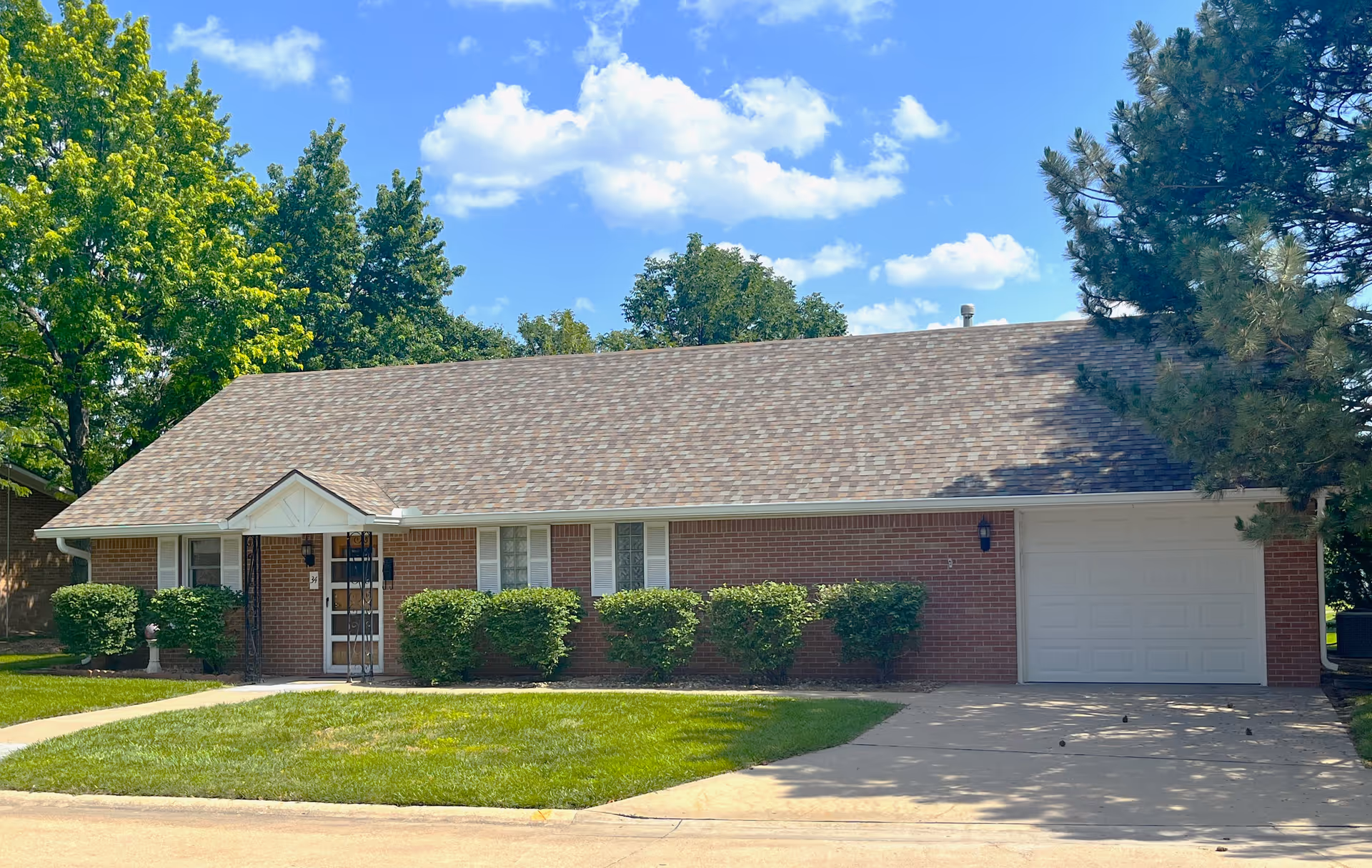 Single-story brick house with a gray shingled roof, white garage door, and white-framed windows. The front yard has neatly trimmed bushes and a well-maintained lawn. Trees surround the house under a blue sky with scattered clouds.