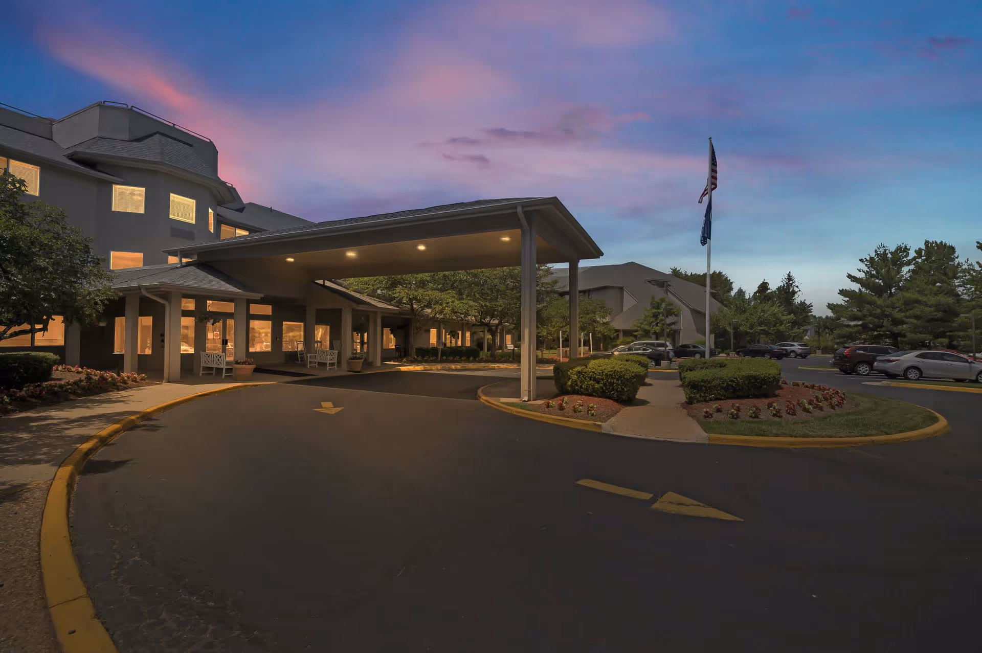 Dusk view of a senior living facility entrance with a covered porte-cochere, illuminated windows, landscaped driveway, parked cars, and an American flag.