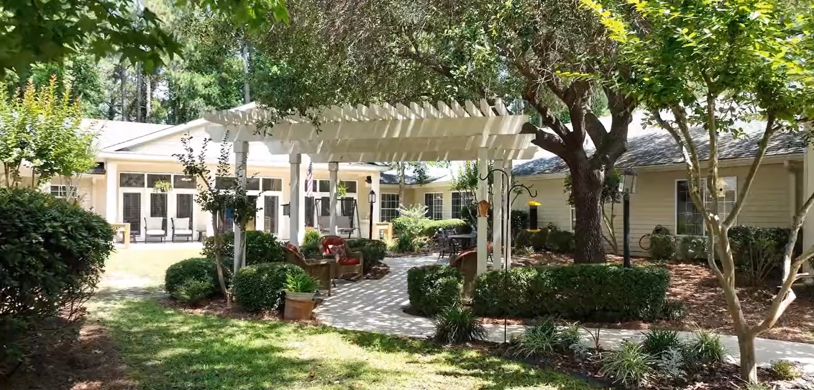 Sunny landscaped courtyard with a white pergola, seating, trees, and the surrounding assisted living buildings.
