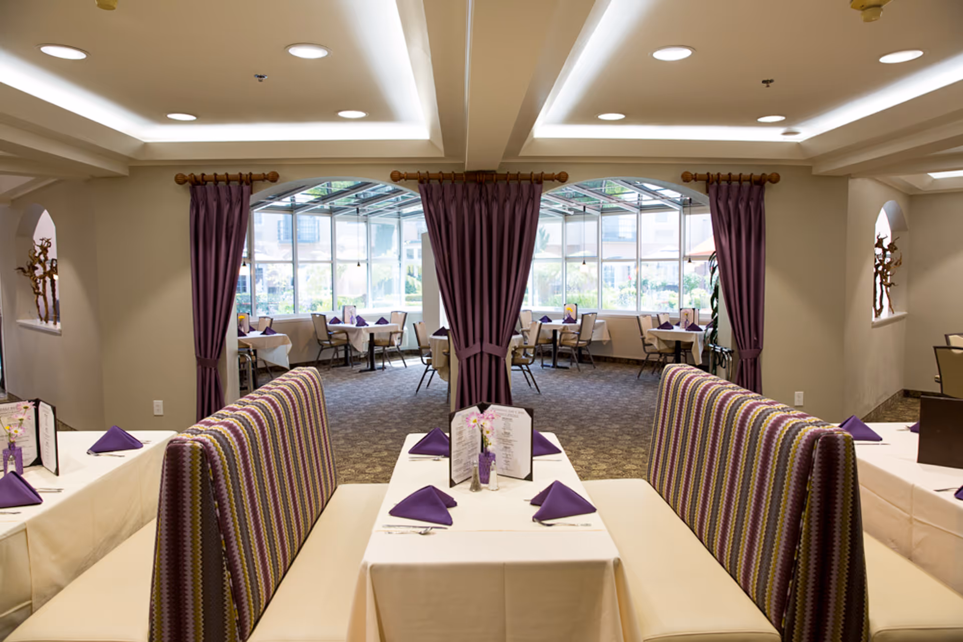 Interior view of a senior living facility dining room with tables covered in white tablecloths, purple napkins, and menus. There are striped booth seats in the foreground and large windows with purple curtains in the background letting in natural light.