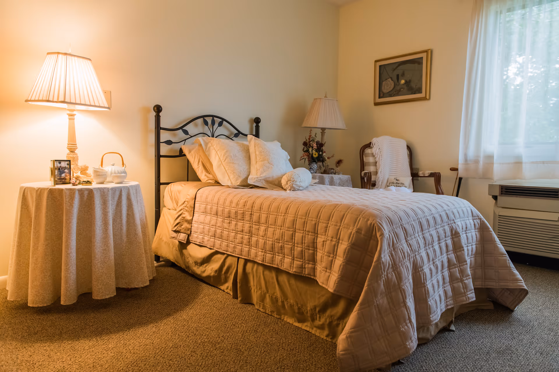 A cozy bedroom with a single bed covered in a beige quilt and multiple pillows. There is a round side table with a lamp, a teapot, and a framed photo on the left side of the bed. On the right side, there is another lamp on a small table, a wooden chair with a white blanket draped over it, a framed picture on the wall, and a window with sheer curtains letting in natural light.